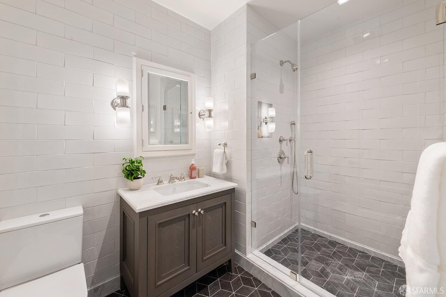 This modern bathroom features a sleek gray vanity with a white marble countertop, complemented by white subway tile walls and a striking dark geometric tile floor. A glass-enclosed walk-in shower with polished chrome fixtures adds a contemporary touch, while wall-mounted sconces provide elegant lighting. The overall aesthetic is clean, sophisticated, and well-appointed, making it an attractive feature for a home listing.