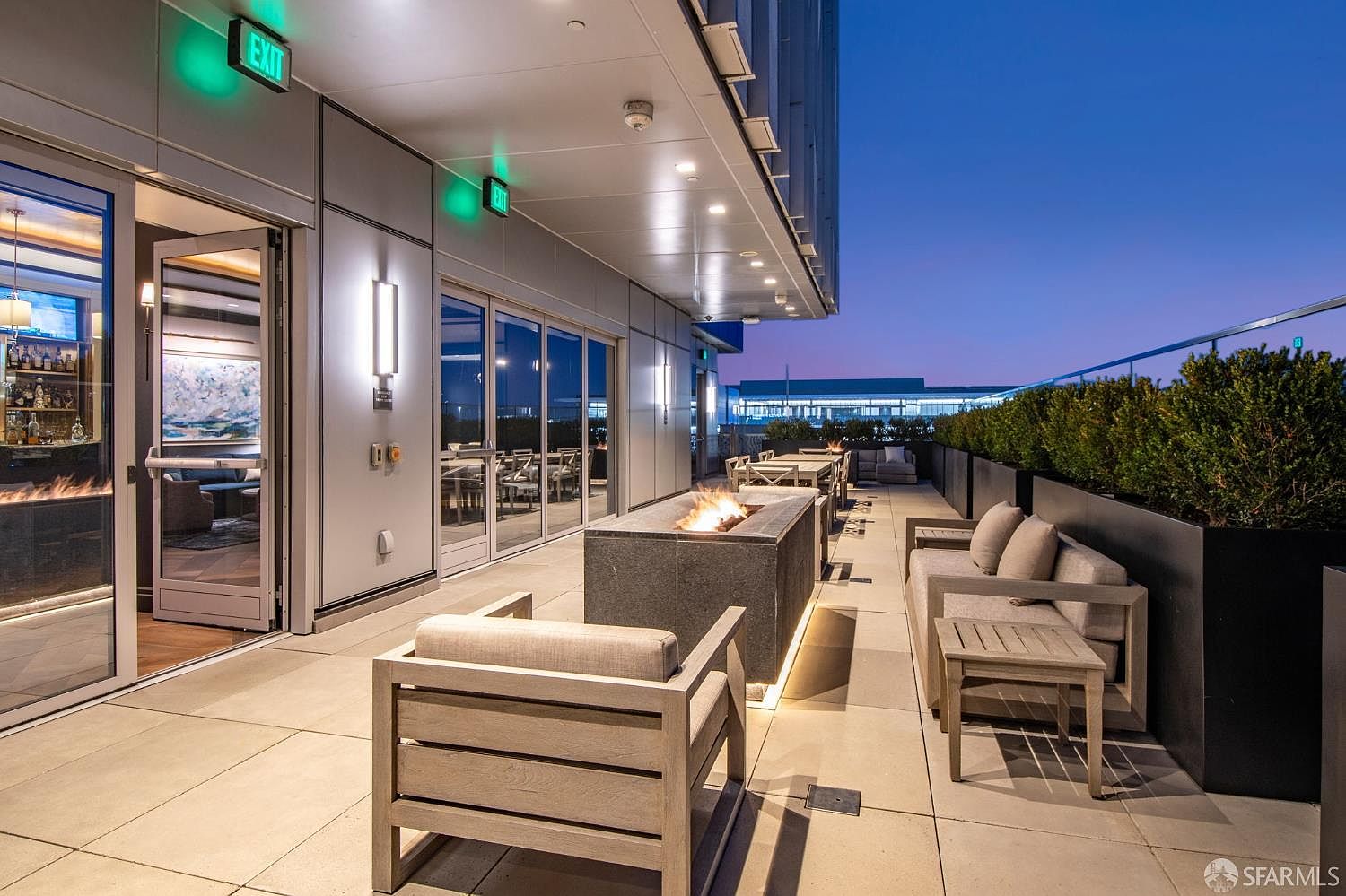 This modern rooftop terrace features a sleek fire pit as a central focal point, surrounded by comfortable lounge seating and contemporary outdoor furniture. The space is framed by a clean, minimalist aesthetic with glass railings and integrated lighting, offering a sophisticated atmosphere for evening relaxation. The perspective captures the seamless transition between the indoor lounge area and the expansive outdoor deck, highlighting the property's premium amenities.