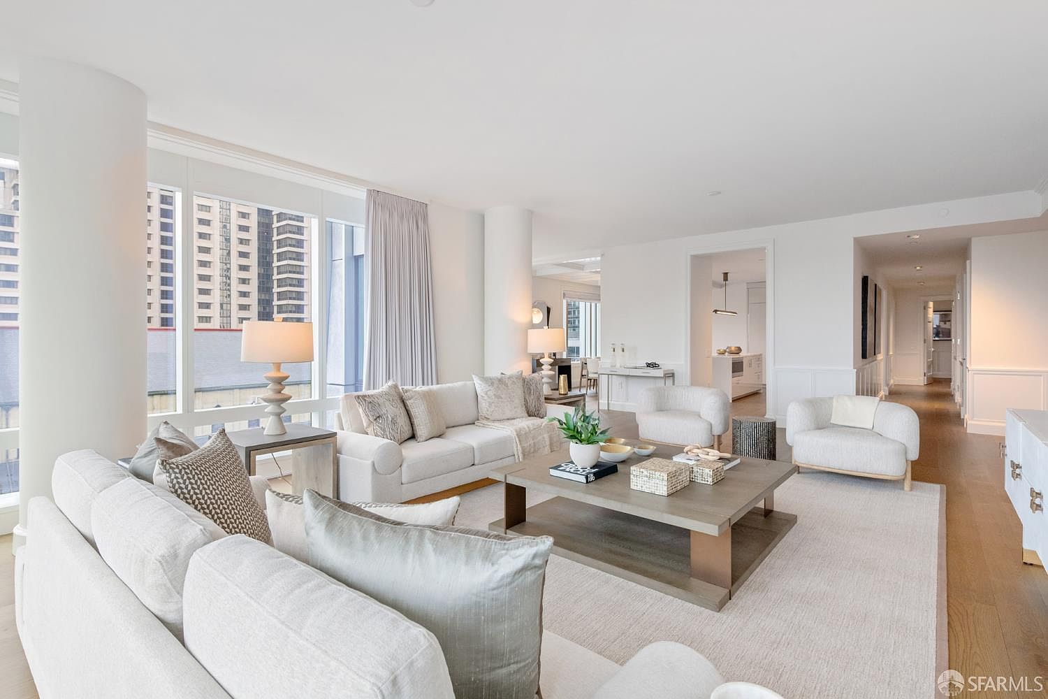 This bright and airy living room features a sophisticated, neutral color palette with plush white sofas, textured throw pillows, and a large, low-profile wooden coffee table. Large floor-to-ceiling windows flood the space with natural light, offering a view of the city skyline, while the open-concept layout seamlessly transitions into a hallway leading to other areas of the home. The overall aesthetic is modern, elegant, and inviting, perfect for high-end urban living.
