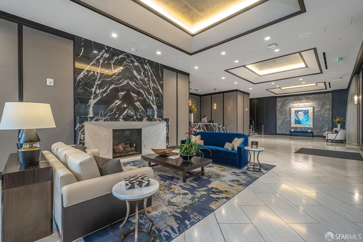 This sophisticated lobby or lounge area features a striking floor-to-ceiling black marble fireplace as its focal point, complemented by a plush blue velvet sofa and cream-colored seating. The space is defined by high ceilings with recessed lighting, polished marble floors, and a large, abstract area rug that ties the seating arrangement together. The overall aesthetic is one of modern luxury and professional elegance, suitable for a high-end residential building or hotel.