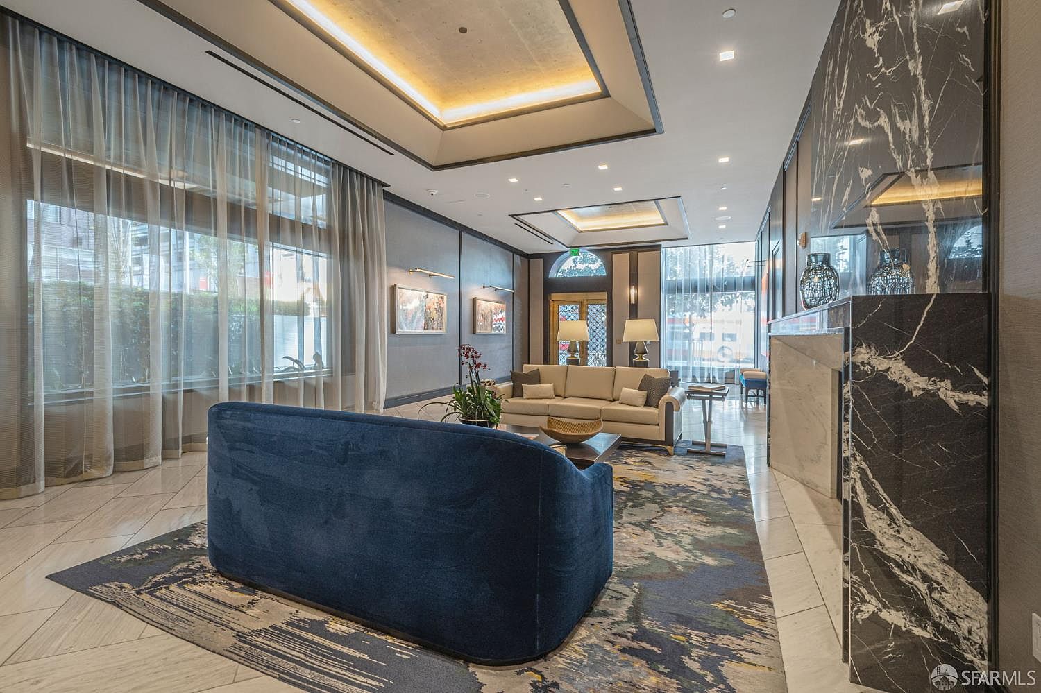 This elegant, high-ceilinged lobby or lounge area features a sophisticated blend of modern and classic design elements, including a striking black marble fireplace and a plush navy blue sofa. The space is anchored by a large, abstract-patterned area rug and illuminated by recessed cove lighting, creating a luxurious and welcoming atmosphere. Large floor-to-ceiling windows draped in sheer curtains allow for soft, natural light to filter into the room, highlighting the polished tile flooring.