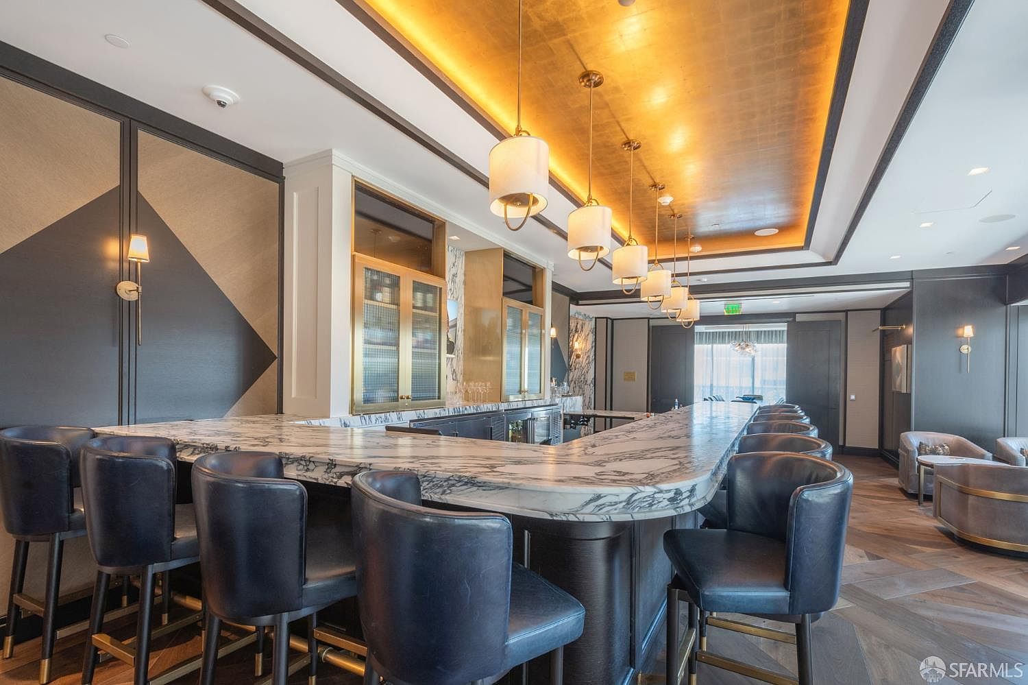 This sophisticated bar area serves as a luxurious community amenity, featuring a long, curved marble countertop with dark leather bar stools. The space is highlighted by a stunning gold-leaf ceiling with elegant pendant lighting and dark, geometric wall paneling. The overall atmosphere is upscale and refined, perfect for social gatherings in a high-end residential building.
