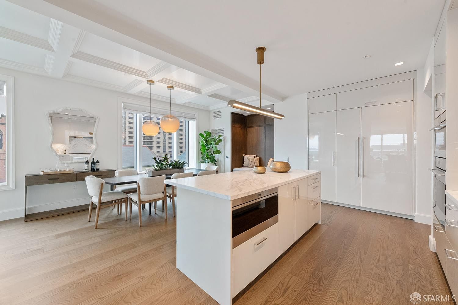 This bright and modern kitchen features a large marble-topped island with integrated appliances, sleek white cabinetry, and high-end finishes. The open-concept space flows seamlessly into a dining area with contemporary pendant lighting and a coffered ceiling, creating an airy and sophisticated atmosphere. Large windows provide ample natural light and a view of the urban surroundings, emphasizing the home's clean, minimalist aesthetic.