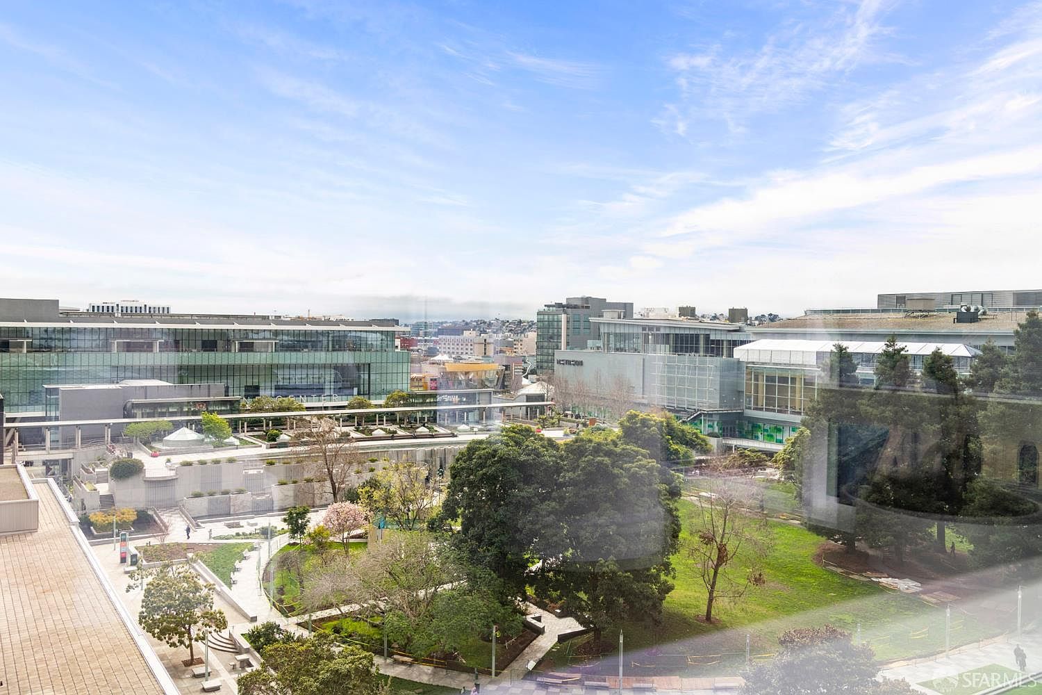 This elevated view captures a serene urban park nestled between modern glass-facade commercial buildings. The scene features lush green lawns, mature trees, and structured walking paths, creating a peaceful oasis within a dense city environment. The perspective is from a high vantage point, likely a window, offering a clear overview of the community green space.