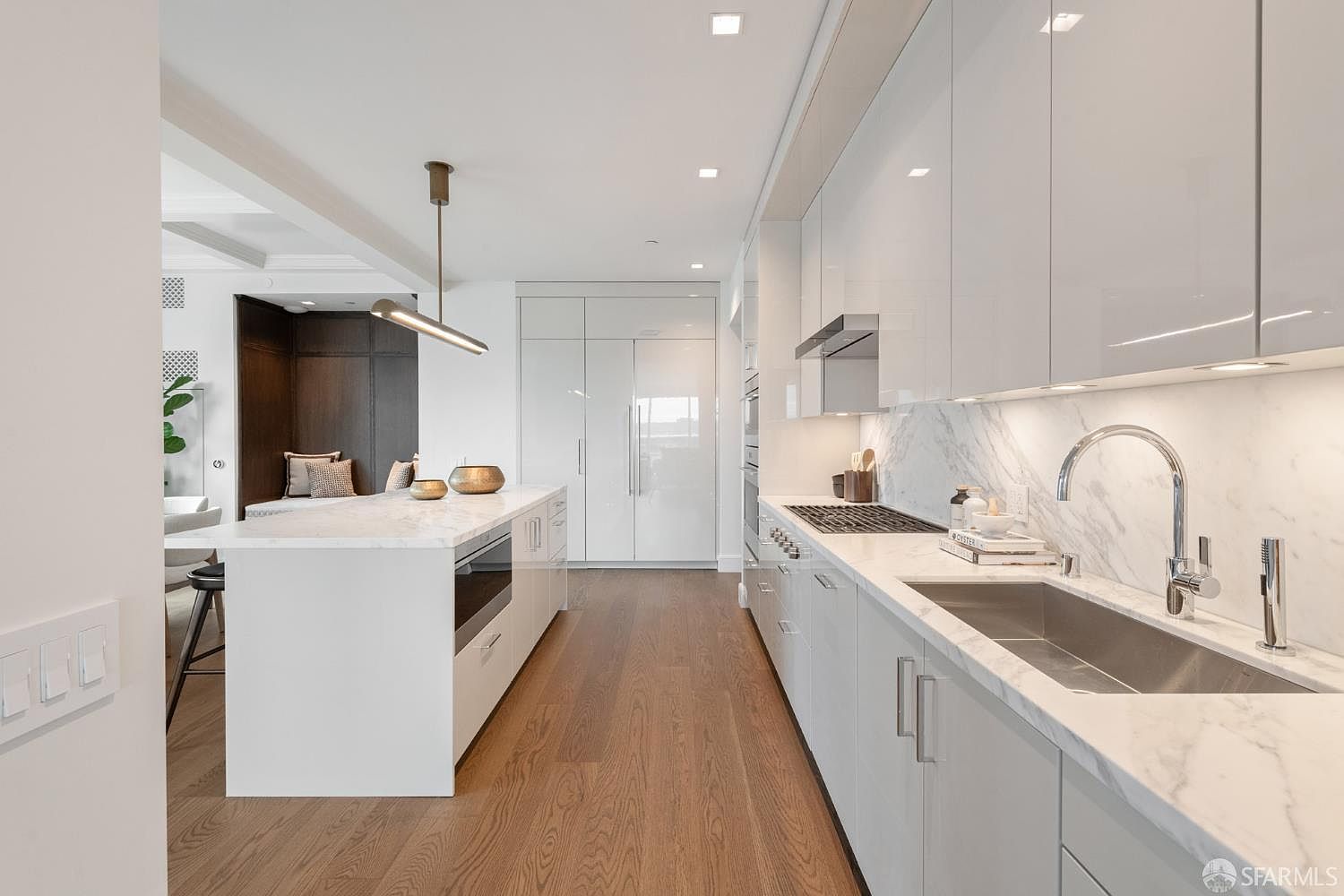 This sleek, modern kitchen features high-gloss white cabinetry, a large marble-topped island, and premium stainless steel appliances. The space is anchored by warm hardwood flooring and illuminated by minimalist pendant lighting, creating a bright and sophisticated atmosphere. The perspective looks down the length of the galley-style layout, highlighting the seamless integration of the kitchen with the adjacent living area.