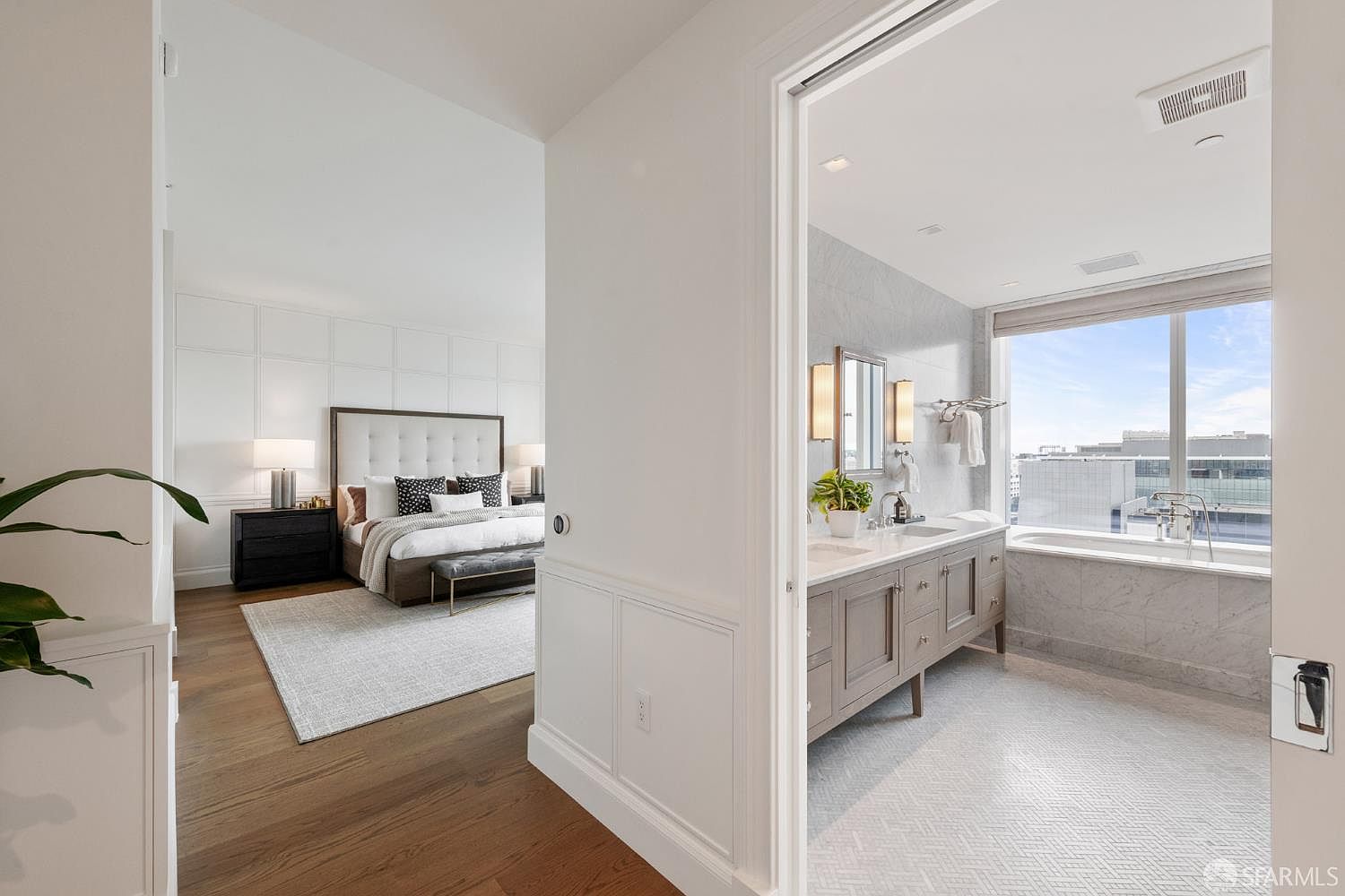 This image captures a sophisticated transition between a primary bedroom and an en-suite bathroom, showcasing a seamless blend of modern luxury and classic architectural detailing. The bedroom features a plush, tufted headboard and a neutral color palette, while the adjacent bathroom boasts a double vanity, marble finishes, and a deep soaking tub positioned beneath a large window with city views. The perspective is framed through a doorway, emphasizing the open, airy flow and high-end finishes of the residence.