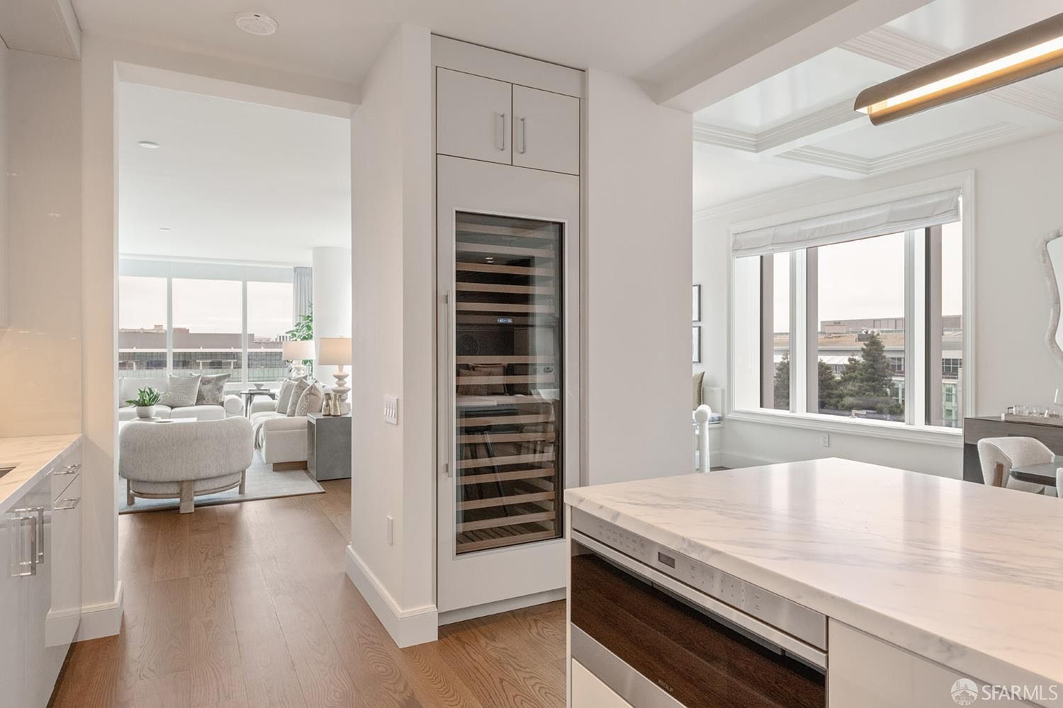 This bright, modern kitchen features a sleek marble-topped island with a built-in microwave drawer and a tall, integrated wine refrigerator set into a white cabinetry wall. The open-concept design provides a clear view into a sunlit living area with hardwood floors, creating a seamless and airy transition between spaces. Large windows throughout the home invite natural light, highlighting the clean lines and sophisticated, contemporary aesthetic.
