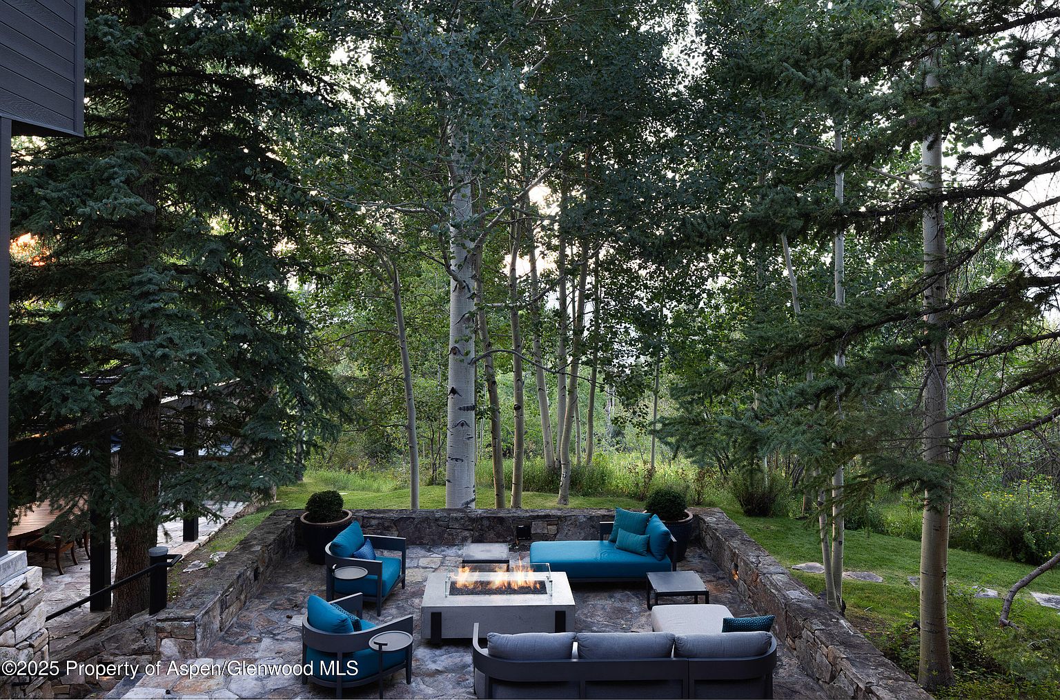 This image showcases a beautifully designed outdoor patio area, complete with comfortable seating arrangements around a modern fire pit. The patio is constructed with stone and surrounded by lush greenery and mature trees, creating a serene and private atmosphere. The space is perfect for entertaining or relaxing outdoors.