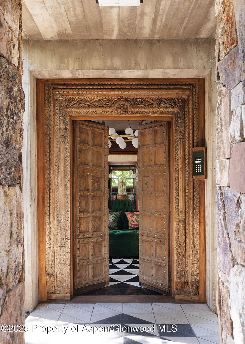 The image showcases an ornate entryway with large, intricately carved wooden doors slightly ajar, revealing a glimpse of the interior. The doorway is framed by stone walls, adding a rustic yet grand feel. Inside, a black and white checkered floor and a plush green seating area are visible, hinting at a stylish and inviting interior space.