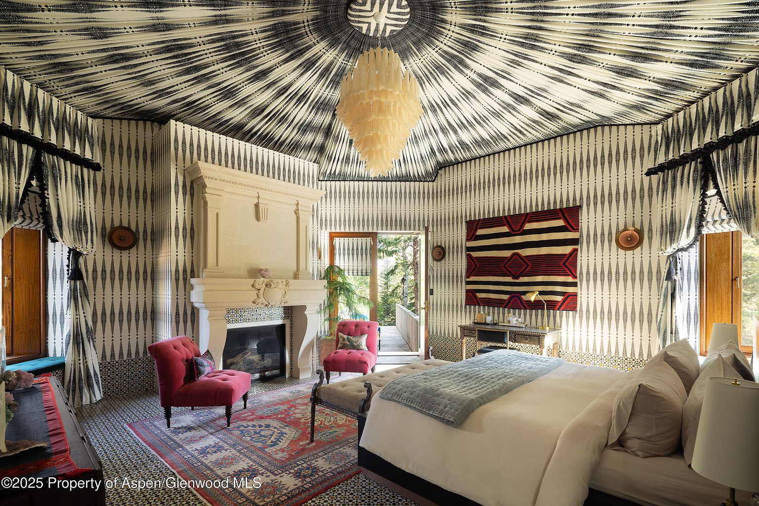 This is a primary bedroom featuring a unique design with patterned wallpaper and ceiling, creating a visually striking space. The room includes a fireplace, two armchairs, a bed with a blue throw, and a decorative chandelier. The overall impression is luxurious and eclectic, with a focus on bold patterns and textures.