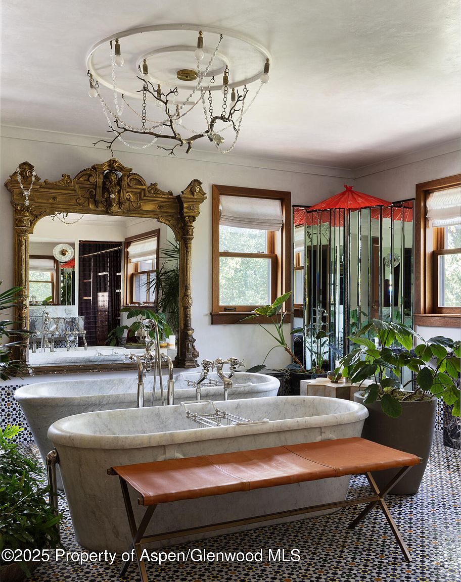 This is a luxurious primary bathroom featuring two marble freestanding bathtubs, each with elegant chrome fixtures. A large, ornate gold mirror reflects the space, enhancing the sense of grandeur. The room is decorated with lush greenery and a unique chandelier, adding a touch of nature and artistry. A leather bench sits in front of one of the tubs. The floor is tiled with a geometric pattern.