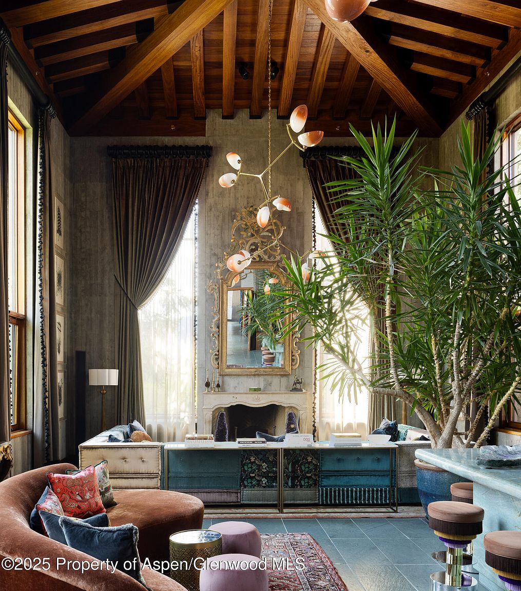 This is an interior shot of a luxurious living room featuring a curved sofa with decorative pillows, a unique chandelier, and a large ornate mirror above a fireplace. The room is decorated with a mix of textures and patterns, including velvet curtains and a patterned rug, creating a warm and inviting atmosphere. Large plants add a touch of nature to the space.