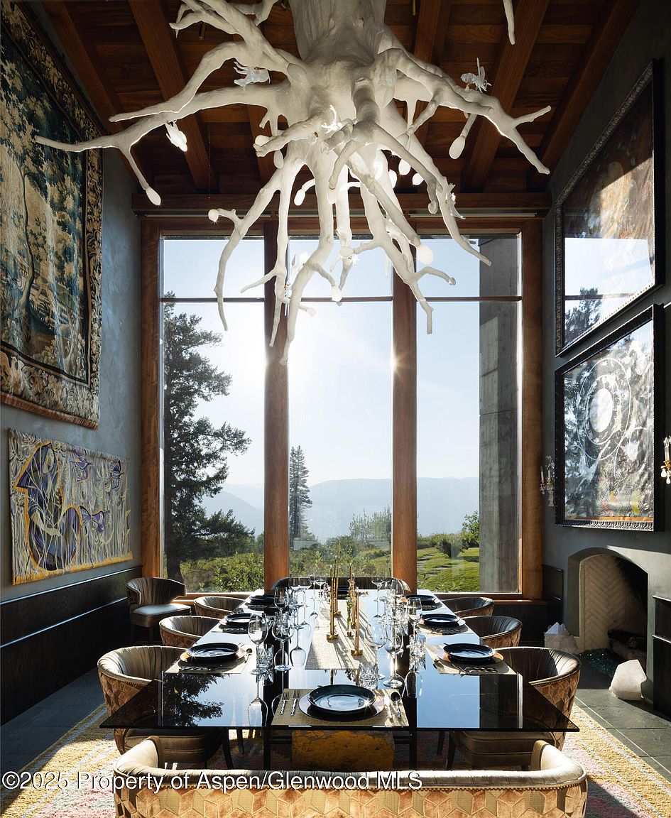 This is an interior shot of a dining room featuring a long, dark, reflective dining table set for a formal meal. Large windows offer a stunning view of the landscape, and a unique, sculptural chandelier hangs above the table, serving as a focal point. Artwork adorns the walls, and the room exudes a sense of luxury and sophistication.