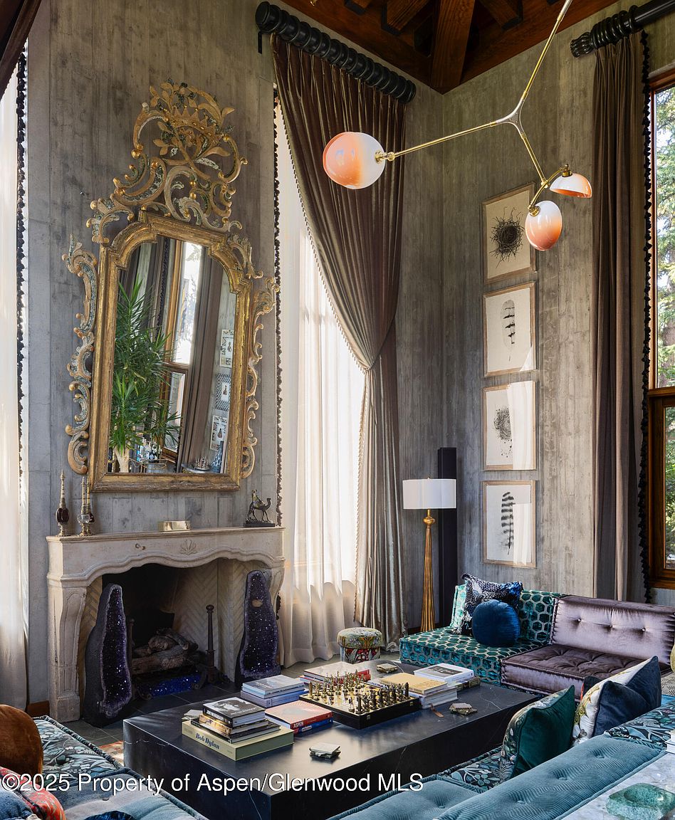 This living room features a grand, ornate mirror above a stone fireplace, flanked by amethyst geodes. The room is decorated with eclectic furniture, including patterned sofas and a coffee table adorned with books and a chess set. Tall windows with heavy drapes provide natural light, complemented by a modern chandelier, creating a luxurious and artistic atmosphere.