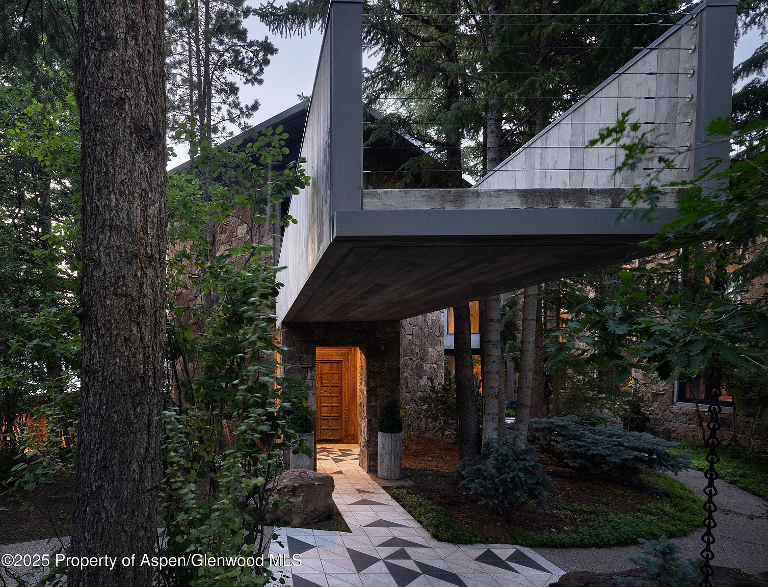 This image showcases the entryway of a modern home, featuring a stone facade and a cantilevered concrete structure above the entrance. A patterned tile walkway leads to a wooden door, with lush greenery and trees surrounding the property. The architectural design is striking and blends seamlessly with the natural landscape.