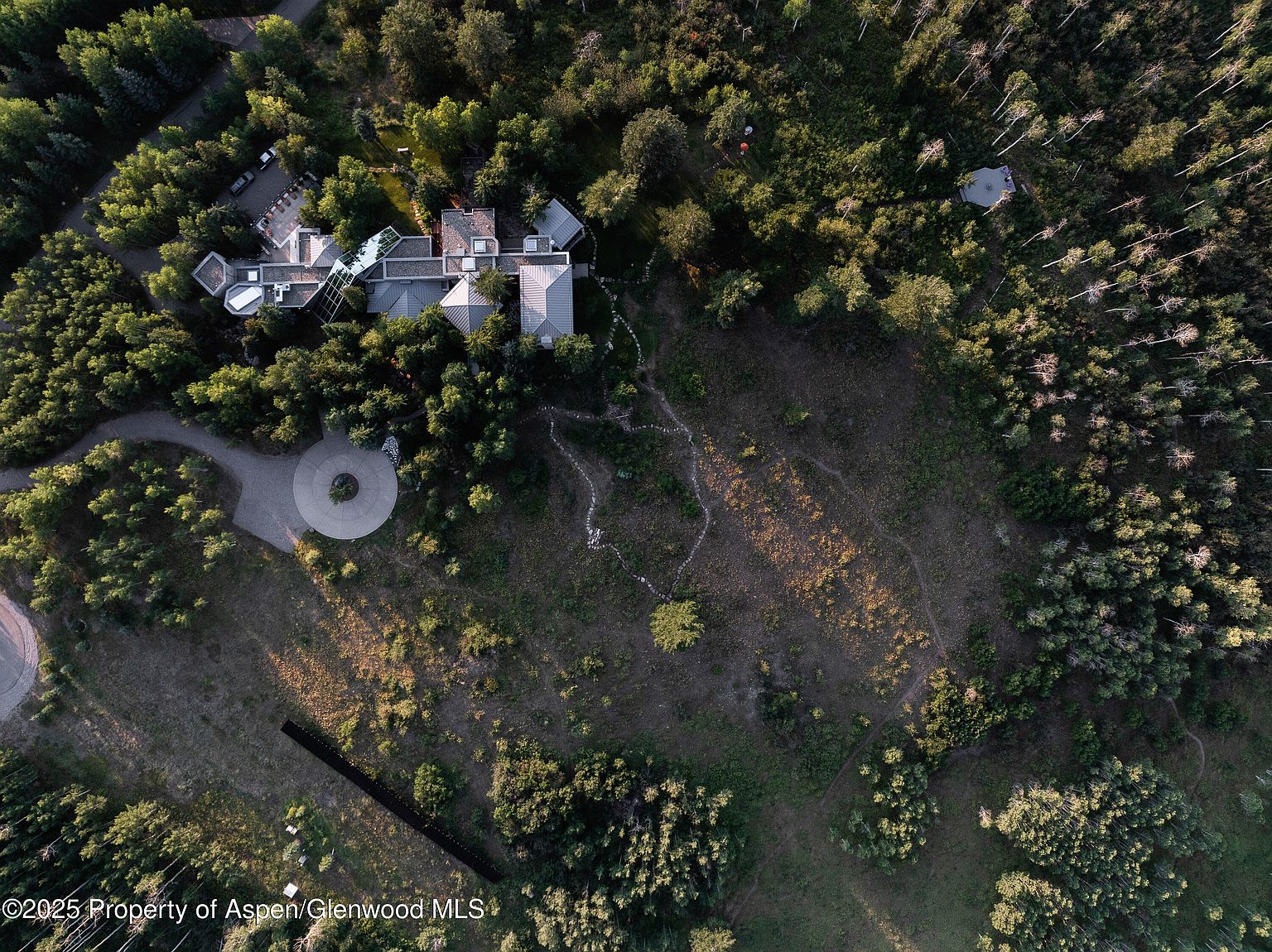 This aerial view showcases a luxurious estate nestled within a dense forest. The property features a sprawling residence with multiple wings and a circular driveway, surrounded by lush greenery and open space. A walking path meanders through the landscape, leading to a secluded area within the trees, highlighting the property's privacy and natural setting.