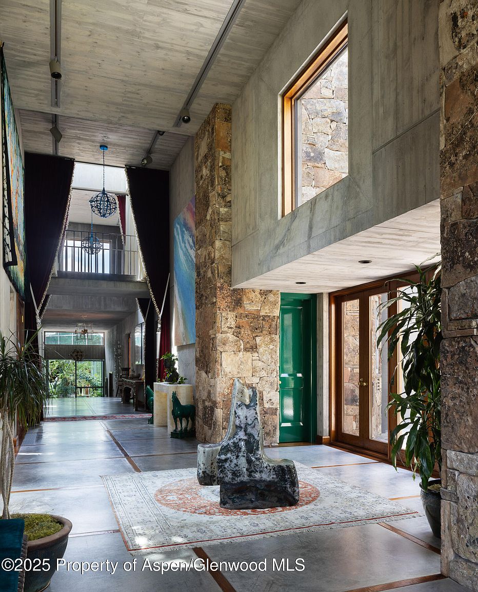 This grand hallway features a striking blend of modern and natural elements. Concrete walls and ceilings contrast with stone accents, while a large area rug anchors the space. A unique sculpture serves as a focal point, and the open layout provides a glimpse of the upper level and outdoor views, creating an airy and inviting atmosphere.