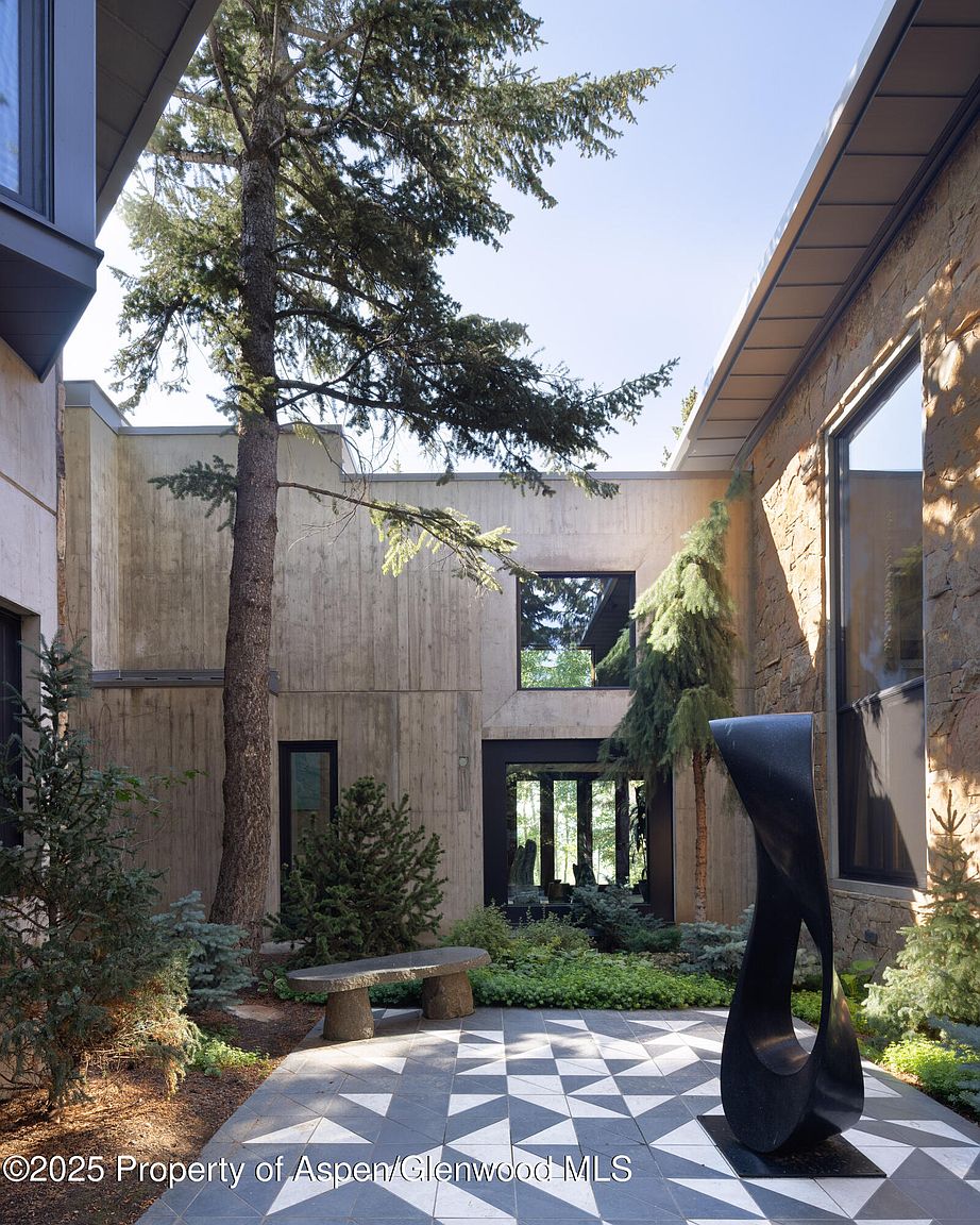 This image showcases a modern courtyard garden, featuring a striking black sculpture as a focal point. The space is paved with a geometric tile pattern and includes a stone bench nestled among lush greenery and trees. The surrounding architecture is a blend of concrete and stone, creating a sophisticated and serene outdoor living area.
