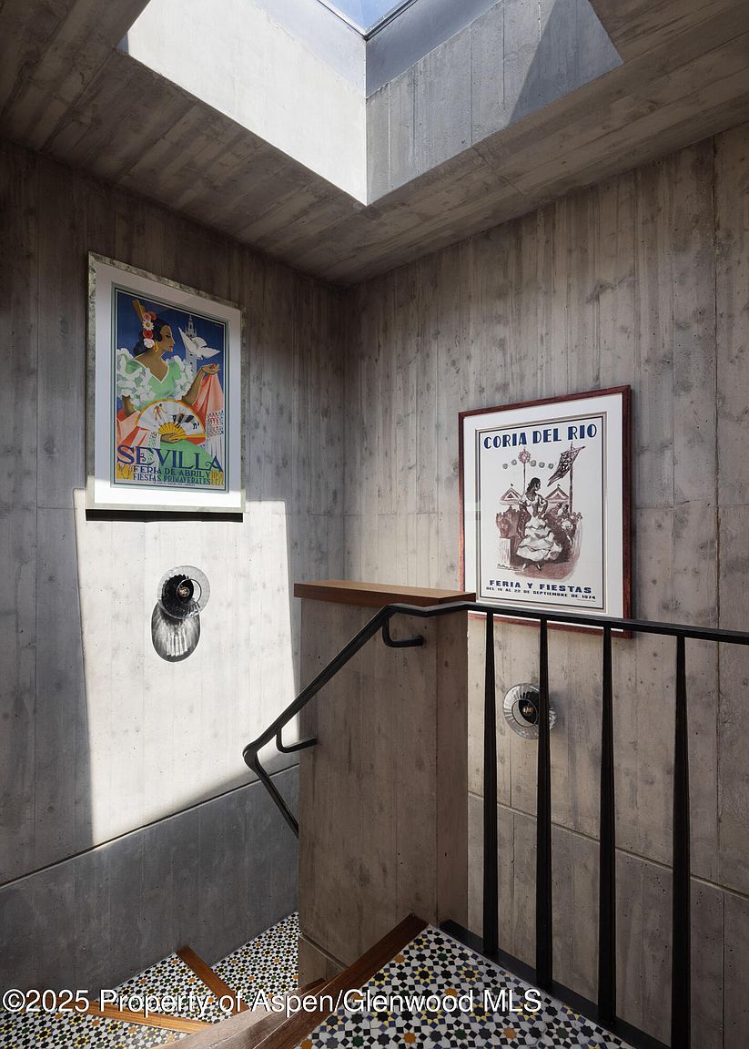 This image showcases a modern stairwell with concrete walls and decorative tile steps. Two framed posters adorn the walls, adding a touch of character to the otherwise minimalist space. A black metal railing provides a sleek contrast to the raw concrete, and a skylight above allows natural light to flood the area.