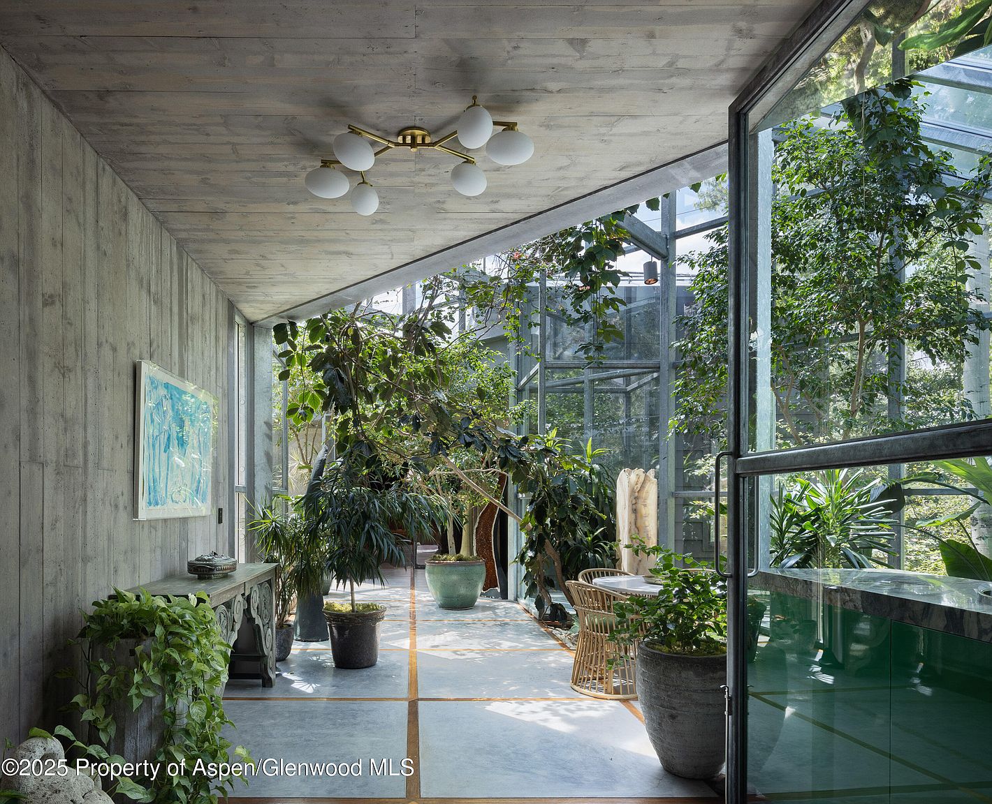 This image showcases a modern sunroom or conservatory with a concrete ceiling and walls, complemented by an abundance of lush greenery and plants. The room features large glass windows and doors, creating a seamless connection with the outdoors. The space is well-lit and airy, offering a tranquil and inviting atmosphere.