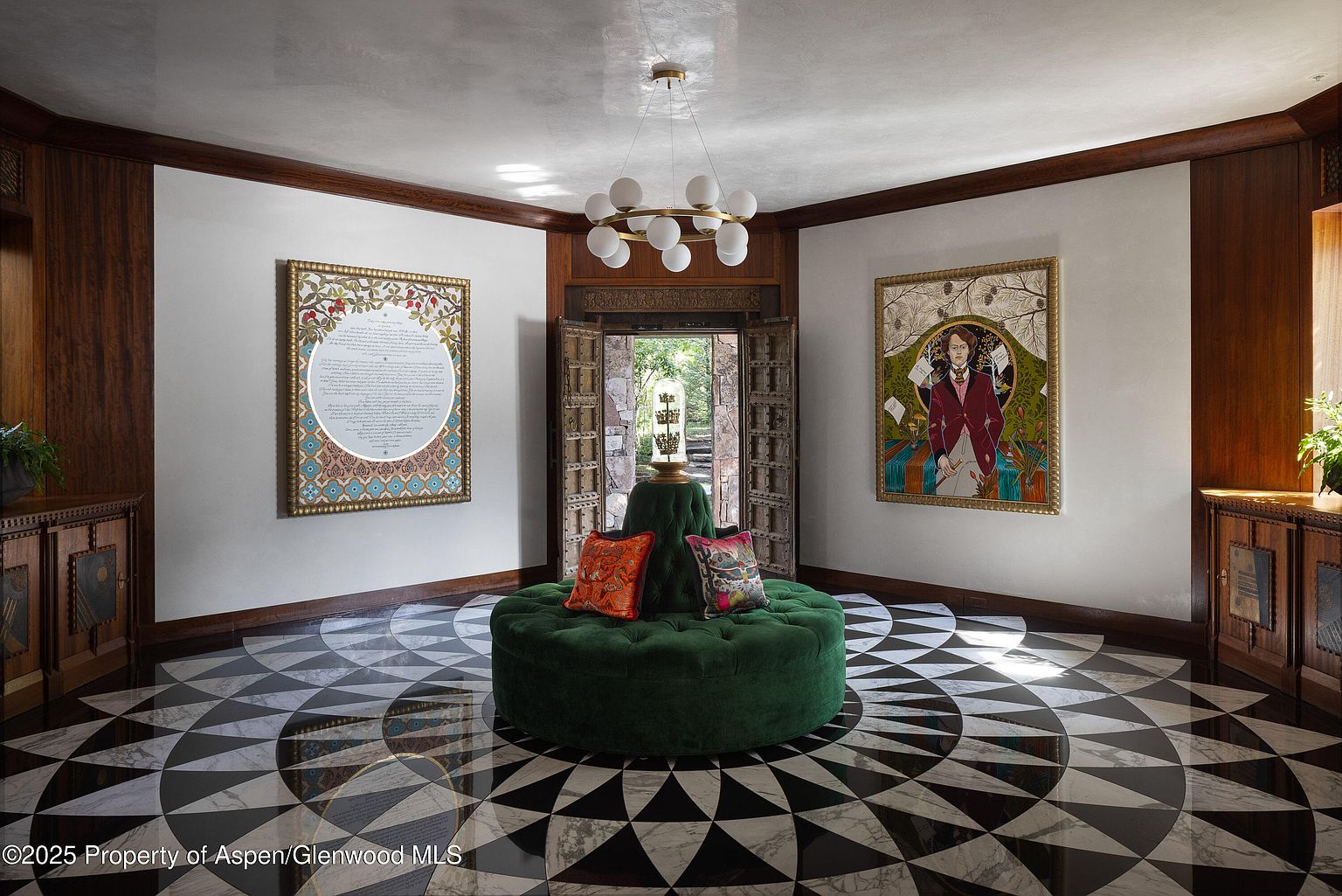 This grand entryway features a striking black and white patterned floor, a large round green ottoman with decorative pillows, and ornate artwork on the walls. The space is illuminated by a modern chandelier, and the dark wood trim adds a touch of traditional elegance. The open doorway provides a glimpse of the exterior, creating a welcoming and luxurious atmosphere.