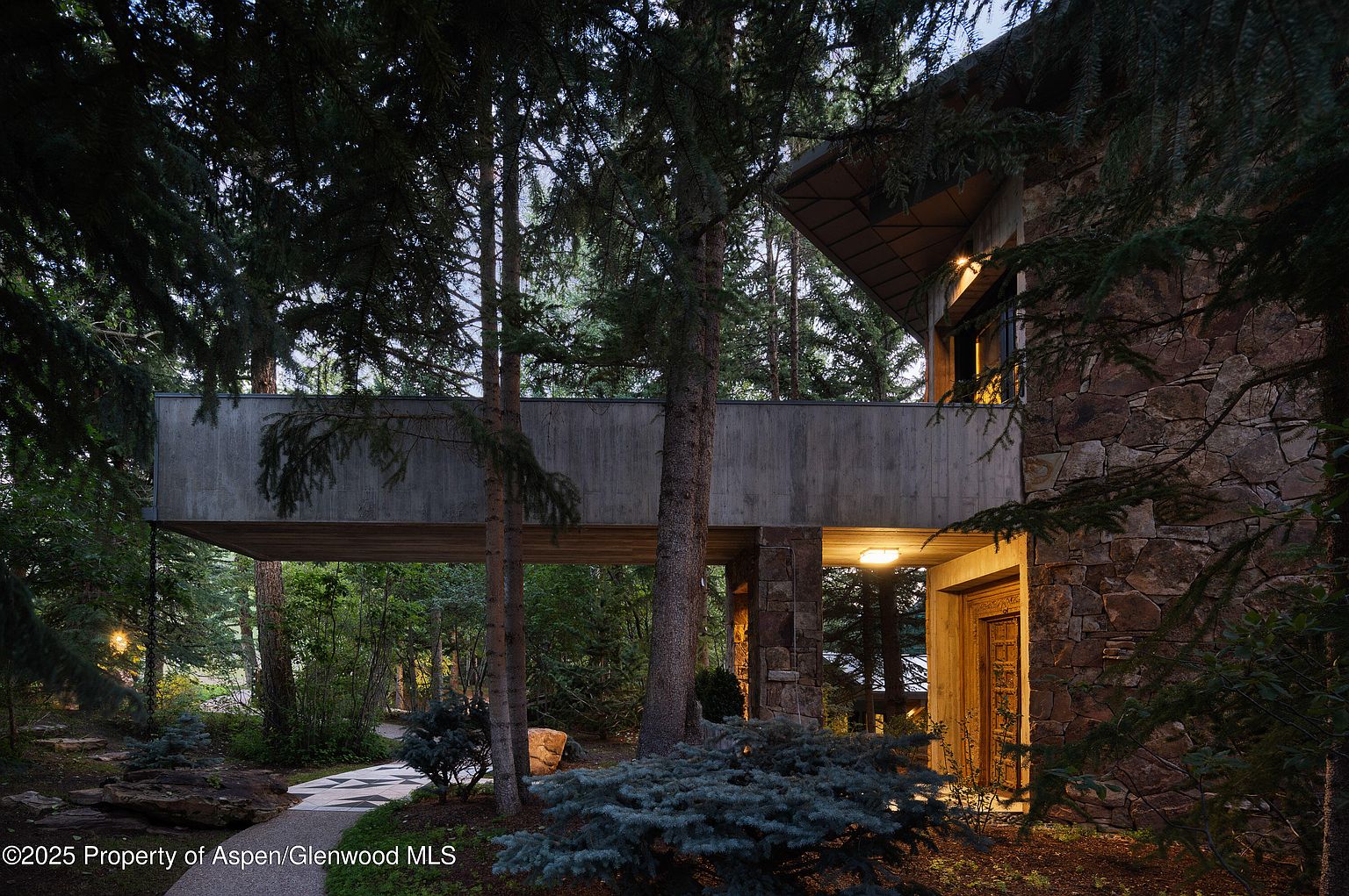 This image showcases the exterior entryway of a unique home, featuring a stone facade and a modern concrete bridge connecting to another part of the house. The entrance is illuminated by warm lighting, highlighting the wooden door and the surrounding landscaping with lush greenery and a patterned walkway. The overall impression is one of sophisticated design integrated with a natural setting.