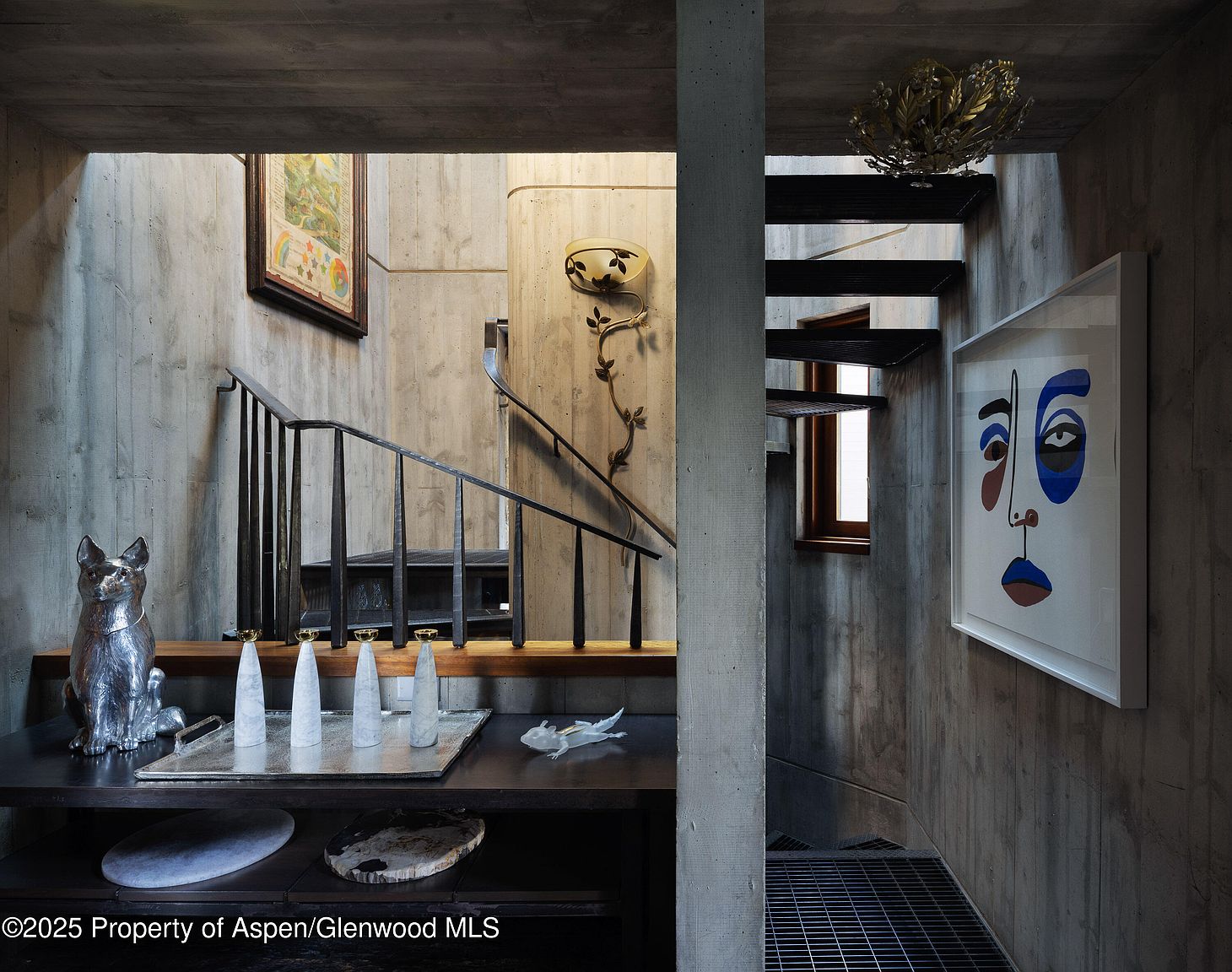 This interior shot showcases a modern hallway and staircase with a raw, industrial aesthetic. The walls and structural elements are primarily exposed concrete, complemented by black metal railings and stair treads. Art pieces and decorative objects add a touch of sophistication to the otherwise minimalist space, creating a unique blend of textures and styles.