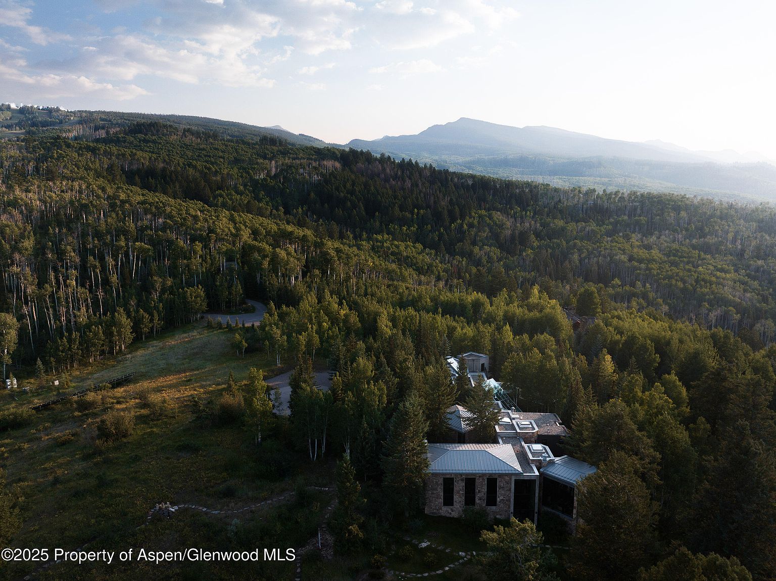 This aerial view showcases a luxurious home nestled amidst a dense forest, offering privacy and stunning natural surroundings. The property features a multi-level structure with a stone facade and a metal roof, blending seamlessly with the landscape. A winding road leads to the house, emphasizing its secluded location and the expansive greenery surrounding it.