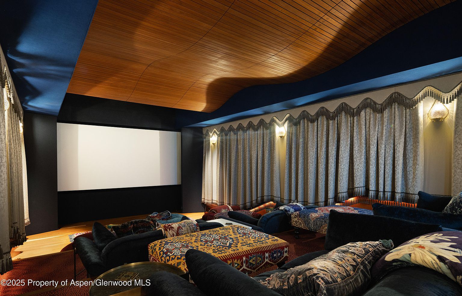 This is an interior shot of a home theater. The room features a large screen, comfortable seating arrangements with couches and floor cushions, and unique architectural details such as a curved wooden ceiling and draped curtains with sconces. The overall impression is one of luxury and comfort, perfect for a cinematic experience.