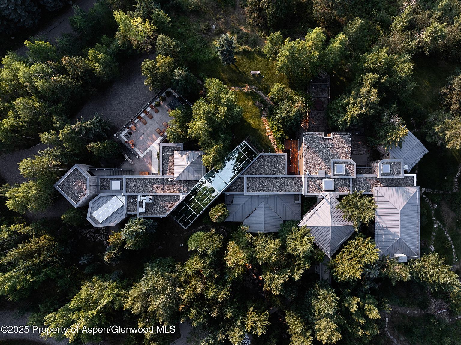 This aerial shot showcases a sprawling, modern residence nestled among lush greenery. The architectural design features a combination of flat and pyramidal roofs, connected by a glass-enclosed walkway. A large patio area with outdoor seating is visible, suggesting ample space for entertaining and relaxation.