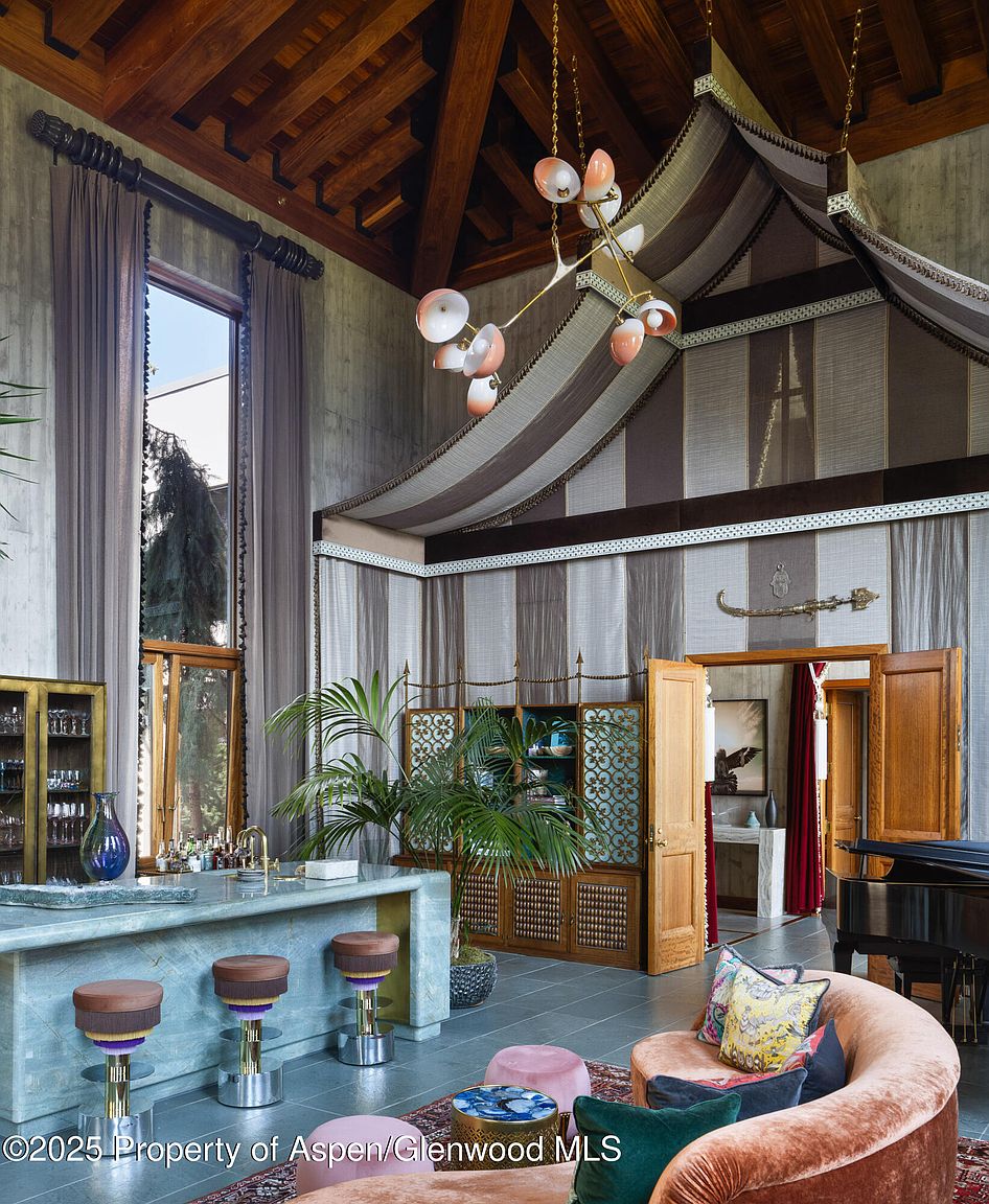 This interior shot showcases a unique living room with a high, exposed-beam ceiling and a custom-designed tented canopy. A marble bar with stools, a grand piano, and a curved sofa create a luxurious and eclectic atmosphere. The room is well-lit by a modern chandelier and natural light from a large window, highlighting the architectural details and upscale furnishings.