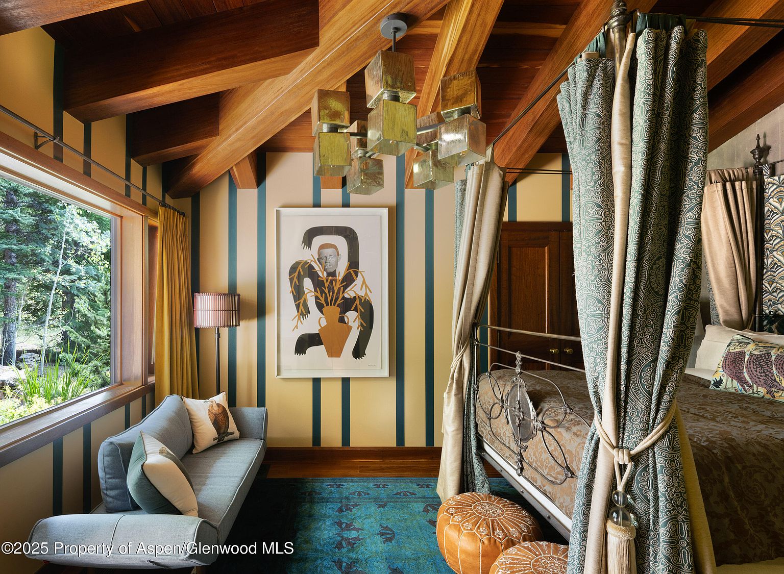 This is a bedroom featuring a four-poster bed with patterned curtains, a blue sofa near a window, and a unique art piece on the striped wall. The room has a cozy and eclectic style, with wooden beams on the ceiling adding a rustic touch. The overall impression is a blend of comfort and artistic flair.
