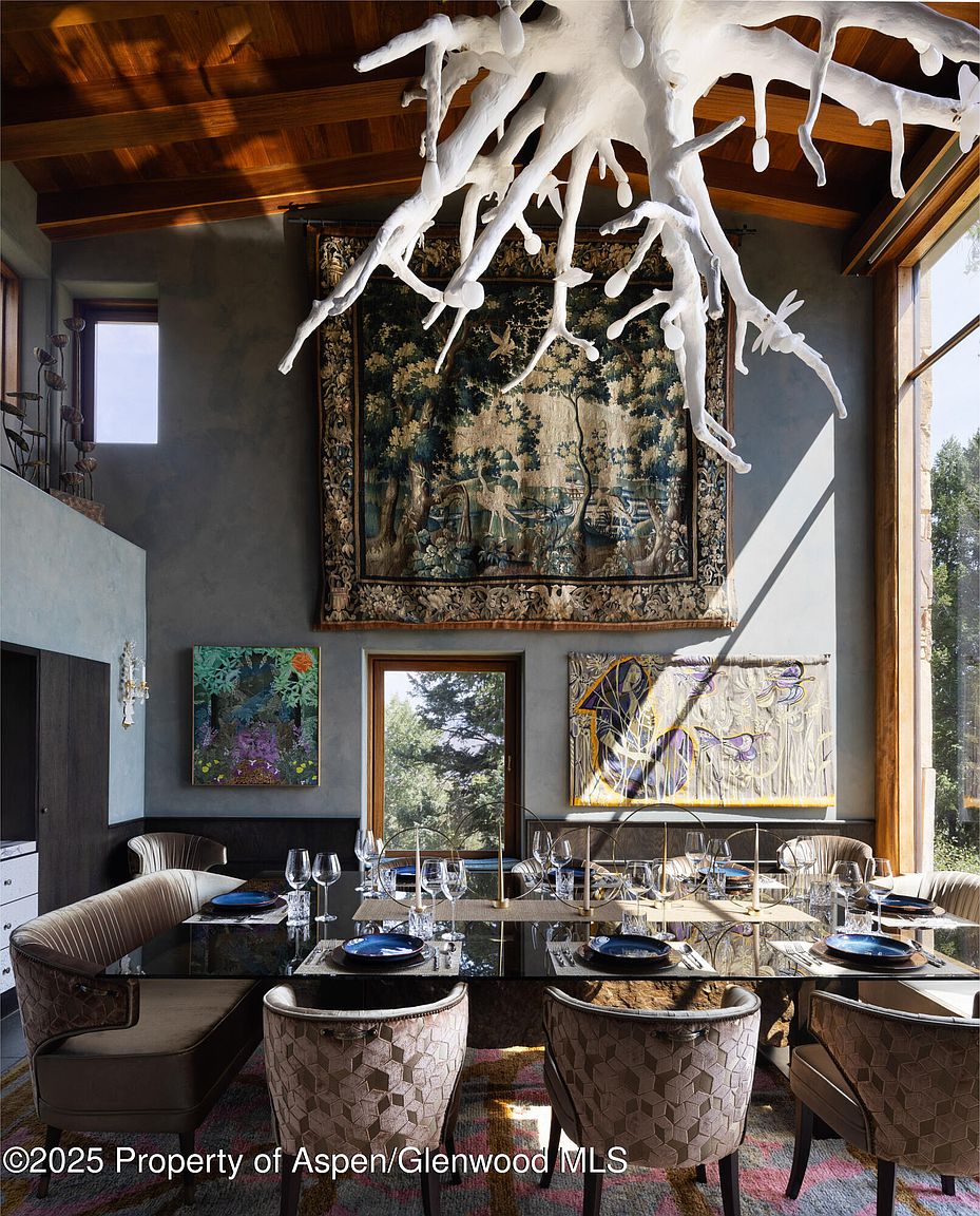 This is an interior shot of a dining room featuring a long, dark glass table set for a formal meal with blue plates and crystal glasses. The room is decorated with a large tapestry and artwork, complemented by a unique, sculptural chandelier. The space is well-lit by natural light streaming through a large window, creating an elegant and inviting atmosphere.
