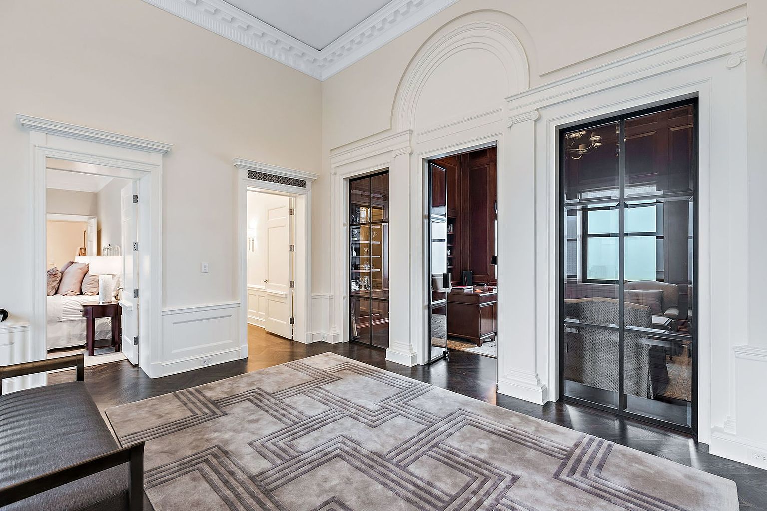 This interior shot showcases a luxurious hallway with dark hardwood floors and a large, patterned area rug. The hallway features multiple doorways, including glass-paneled doors leading to an office or study, and a doorway to a bedroom. The walls are painted in a neutral tone, complemented by white trim and detailed crown molding, creating an elegant and spacious feel.