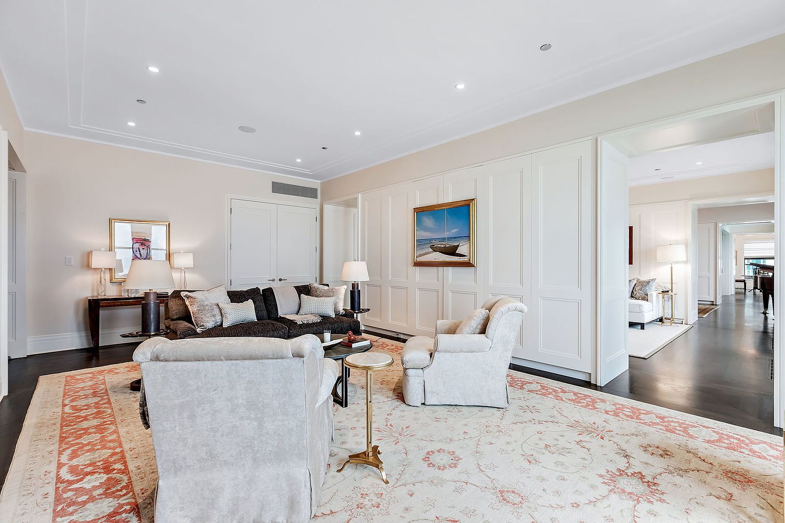 This is an interior shot of a living room featuring a neutral color palette with dark wood floors and a large area rug. The room is furnished with a dark brown sofa, two light-colored armchairs, and a small side table. A painting hangs on the wall, and the room opens into a hallway, creating a sense of spaciousness and flow.