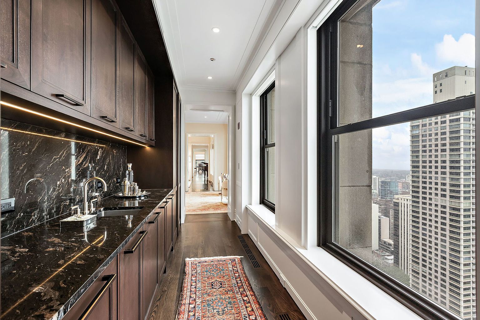 This interior shot showcases a hallway featuring dark wood cabinetry with a marble countertop and sink on one side, and large windows offering a city view on the other. A patterned rug runs along the dark wood floor, leading towards a bright, open space in the distance. The perspective is from a low angle, emphasizing the height and depth of the hallway.