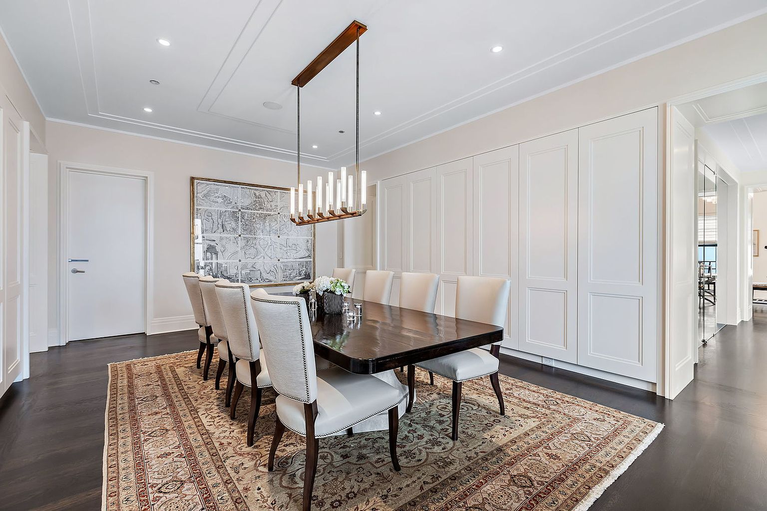 This is an interior shot of a formal dining room. The room features a long, dark wood dining table surrounded by upholstered chairs, a large area rug, and a modern chandelier. The walls are painted in a neutral tone, and there is a large piece of artwork on the wall, creating an elegant and sophisticated atmosphere.