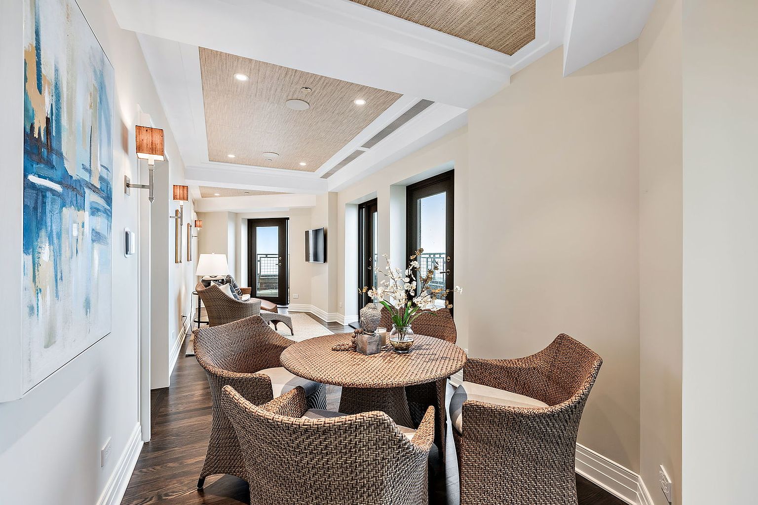 This interior shot showcases a cozy dining area with a round wicker table and four matching chairs, creating an inviting space for meals. The room features a textured ceiling detail, dark hardwood floors, and access to a balcony through dark-framed doors, offering a blend of comfort and sophistication. A large abstract painting adds a touch of modern art to the space.
