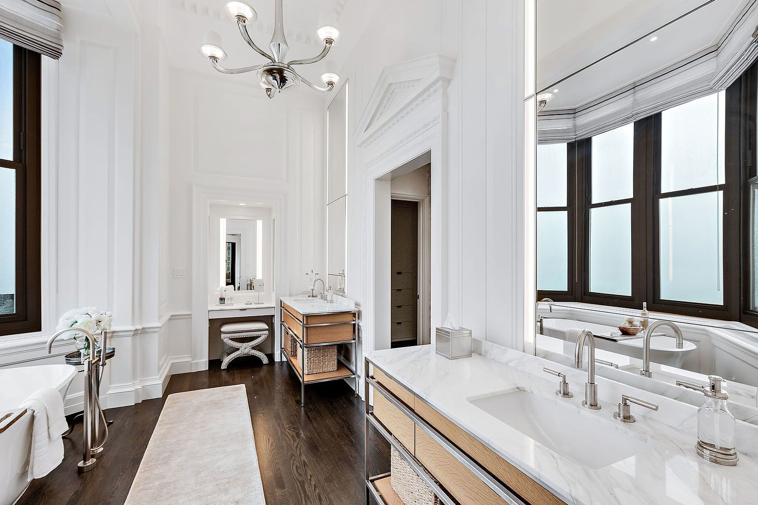 This is a luxurious primary bathroom featuring a double vanity with marble countertops and wooden cabinets. The room is decorated in a classic style with white walls, dark wood floors, and a chandelier. A large mirror reflects the natural light from the window, creating a bright and airy space.
