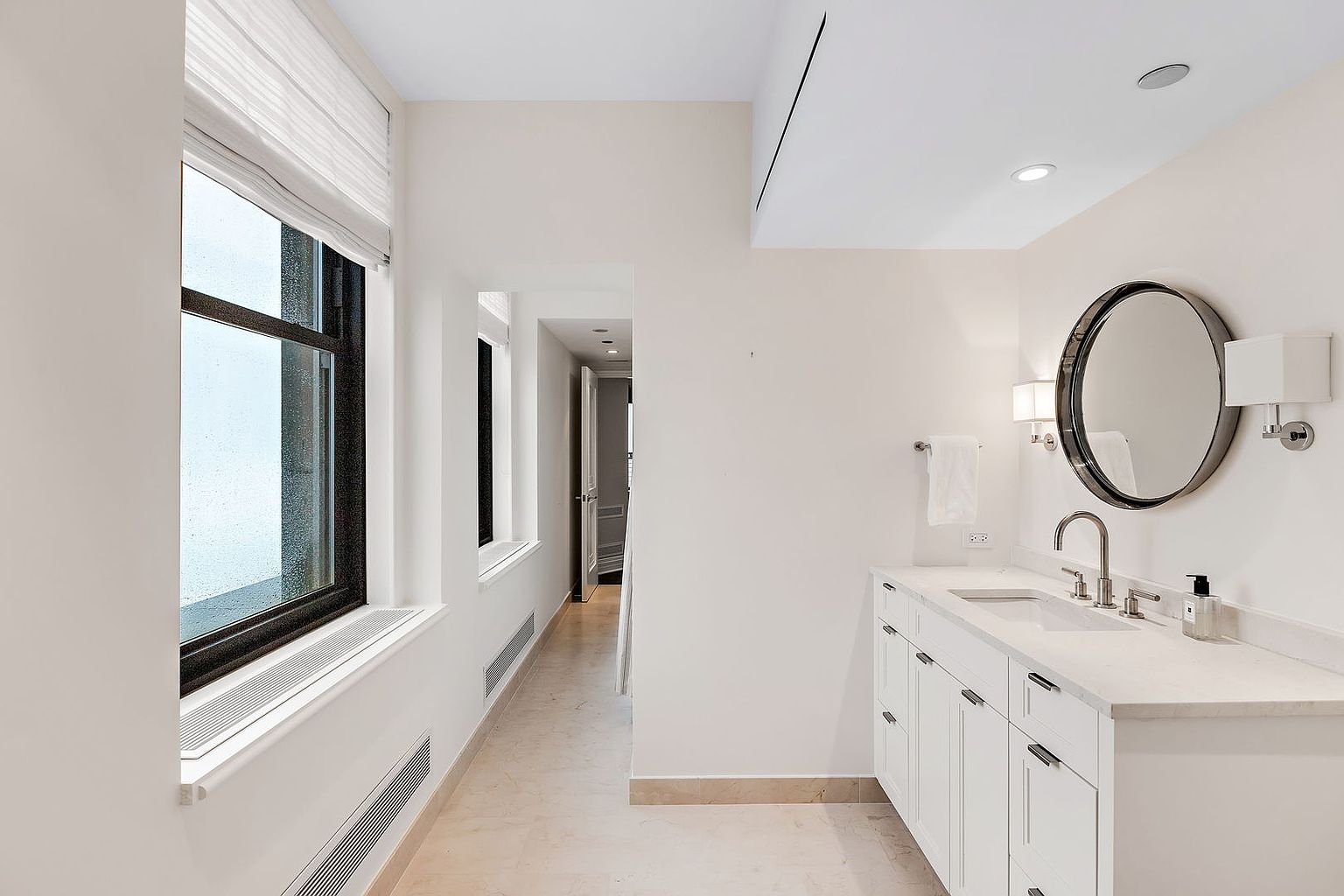 This is a bright and clean bathroom featuring a white vanity with a marble countertop, a round mirror with a dark frame, and modern sconces. A large window provides natural light, and the neutral color palette creates a serene and inviting atmosphere. The perspective is from the doorway, capturing the vanity and the hallway leading further into the home.