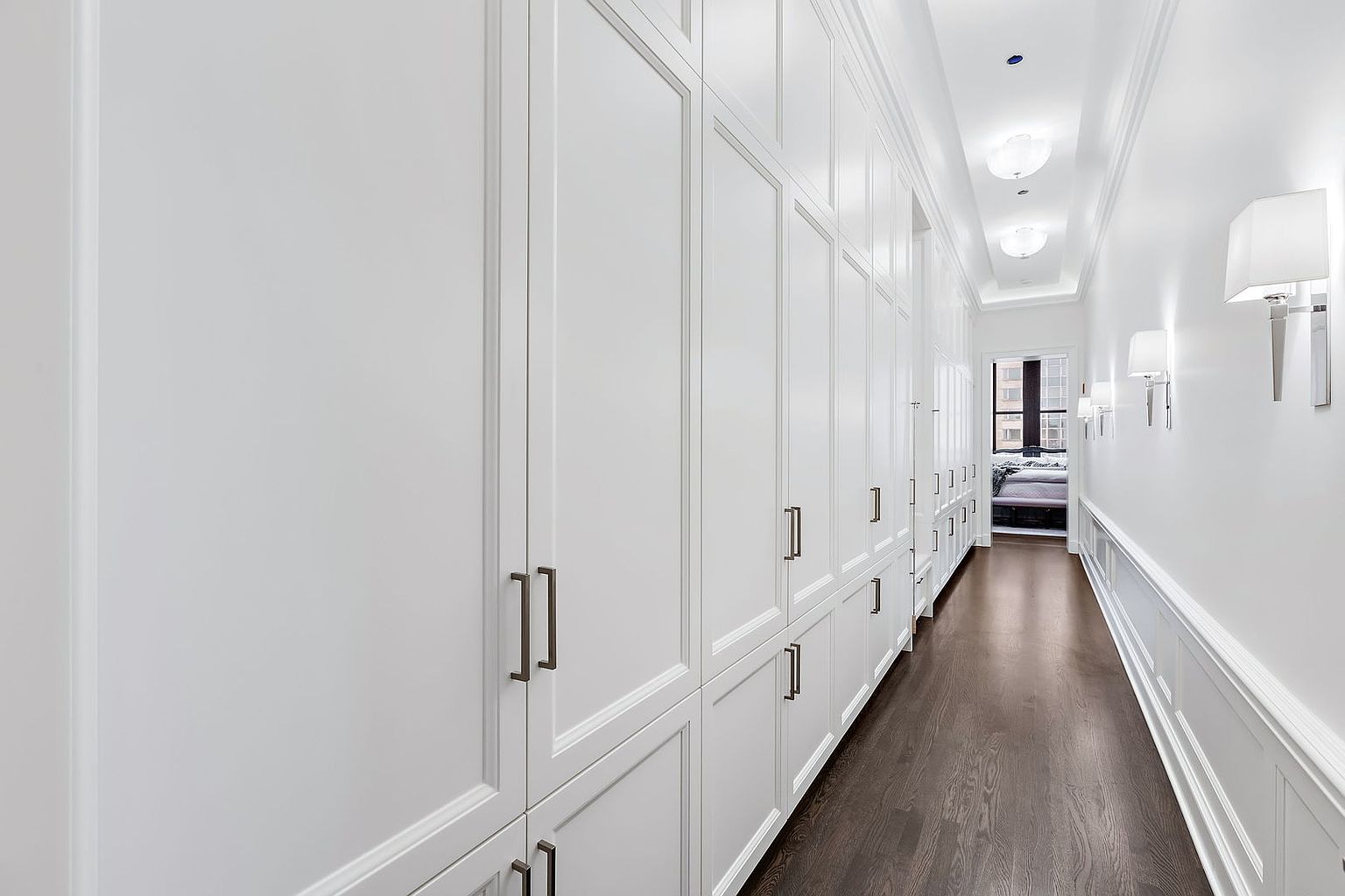 This is a well-lit hallway featuring a long row of built-in white cabinets with sleek, modern handles on one side. The dark hardwood flooring contrasts with the bright white walls and trim, creating a clean and sophisticated look. Wall-mounted sconces provide additional lighting, and a glimpse of a bedroom can be seen at the end of the hallway.