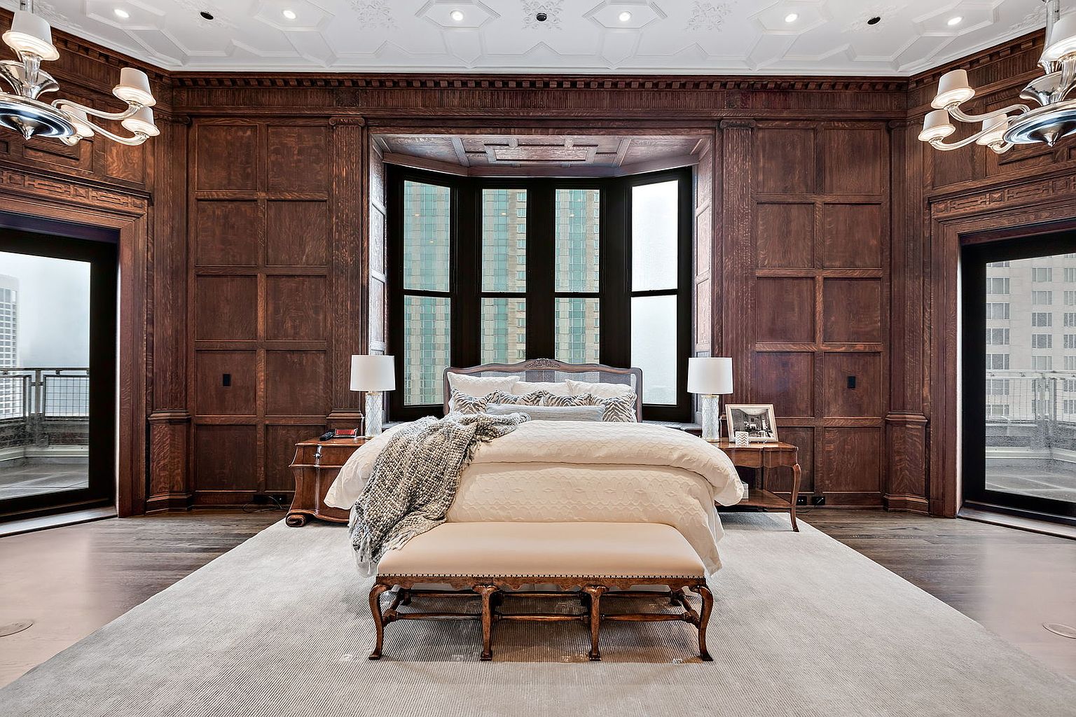 This is a luxurious primary bedroom featuring dark wood paneling, a large bay window, and a neutral color palette. The room is furnished with a king-size bed, side tables with lamps, and a bench at the foot of the bed. The overall impression is one of elegance and sophistication, with a focus on high-end finishes and a comfortable atmosphere.