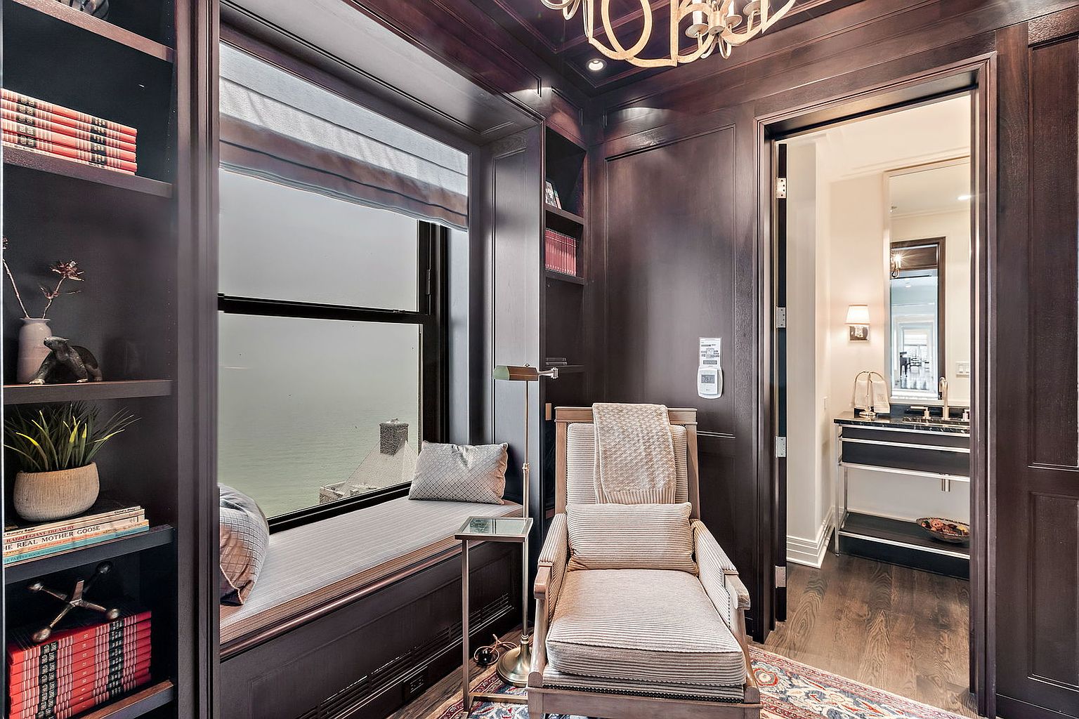 This is an interior shot of a sophisticated home office or study. The room features dark wood paneling, a window seat with a view, a built-in bookcase, and a comfortable armchair. An open doorway leads to a bathroom, suggesting a private and luxurious space.