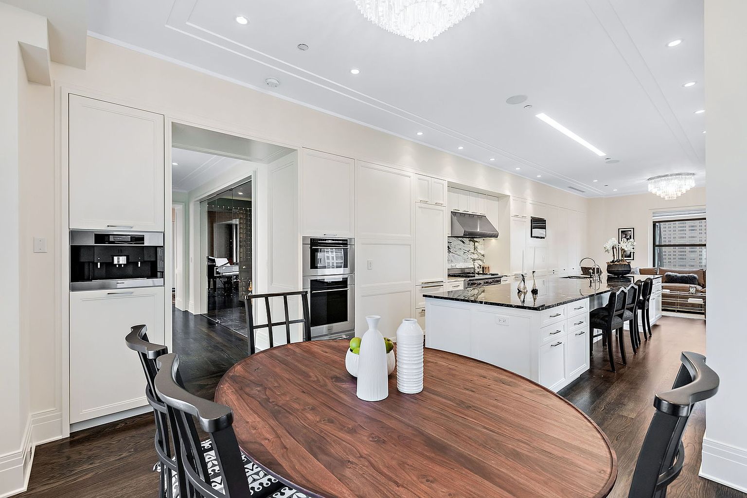 This is a bright and spacious kitchen featuring white cabinetry, stainless steel appliances, and a dark wood dining table. The kitchen island has a black countertop and provides ample space for food preparation. The room is well-lit with recessed lighting and a modern chandelier, creating a clean and elegant atmosphere.