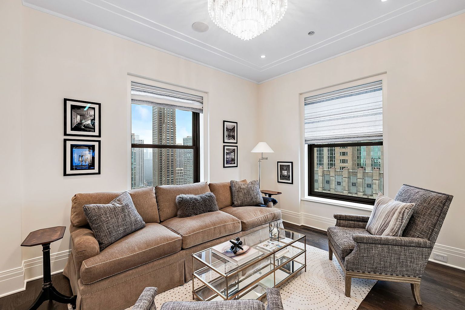 This is a well-lit living room featuring a comfortable brown sofa adorned with decorative pillows, a glass-topped coffee table, and an armchair. Large windows offer city views, enhancing the room's appeal. The space is decorated with framed artwork and a modern chandelier, creating a sophisticated and inviting atmosphere.