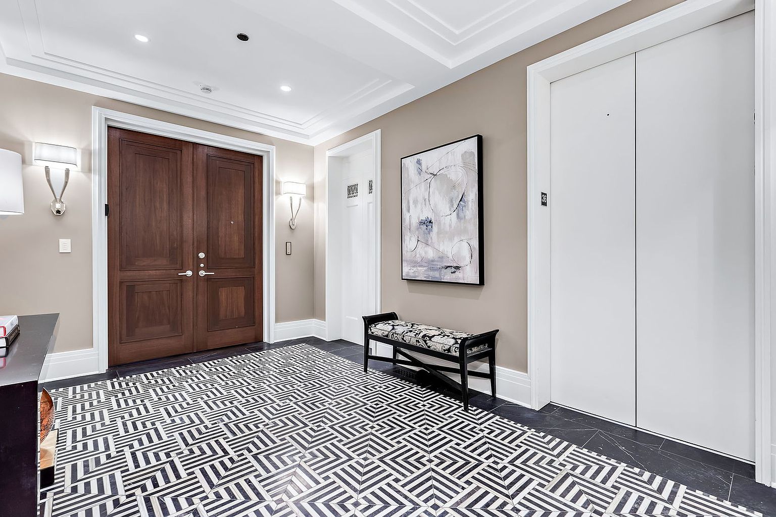 This interior shot showcases a stylish hallway with a striking geometric tile floor and neutral-toned walls. A dark wood double door is visible on the left, complemented by sconce lighting, while a bench sits beneath an abstract painting on the right. The overall impression is one of modern elegance and sophisticated design.