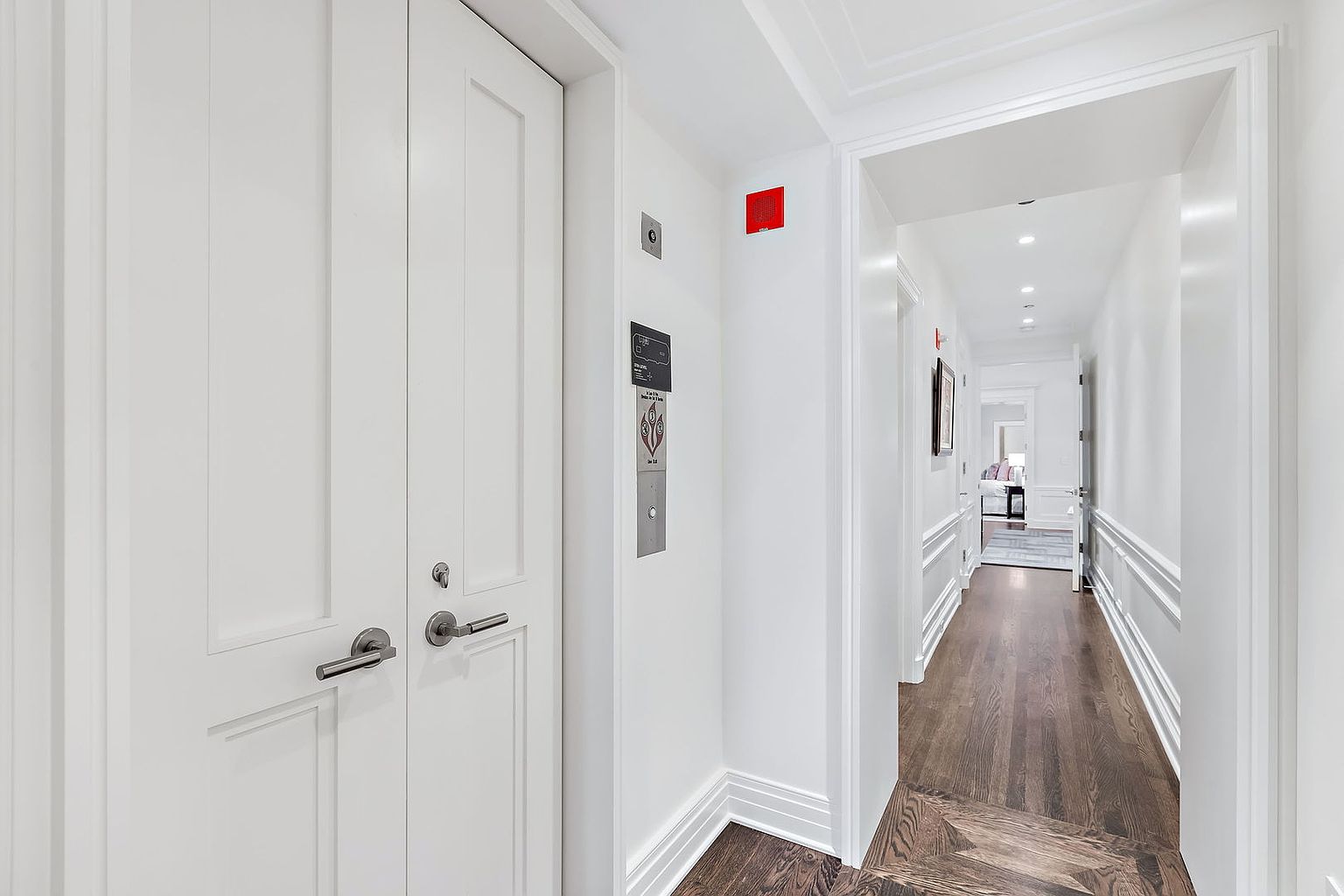 This interior shot showcases a well-lit hallway with dark hardwood floors and white walls. A set of double doors is visible on the left, and an elevator panel is mounted on the wall. The hallway leads to another room, offering a glimpse of a bright interior space, creating a sense of depth and spaciousness.