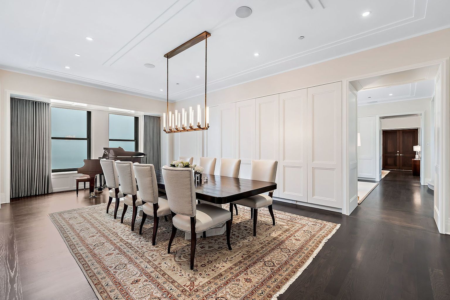 This is an interior shot of a formal dining room. The room features a long, dark wood dining table with upholstered chairs, an ornate area rug, and a modern chandelier. A piano sits near the window, and built-in cabinetry lines one wall, creating an elegant and sophisticated atmosphere.