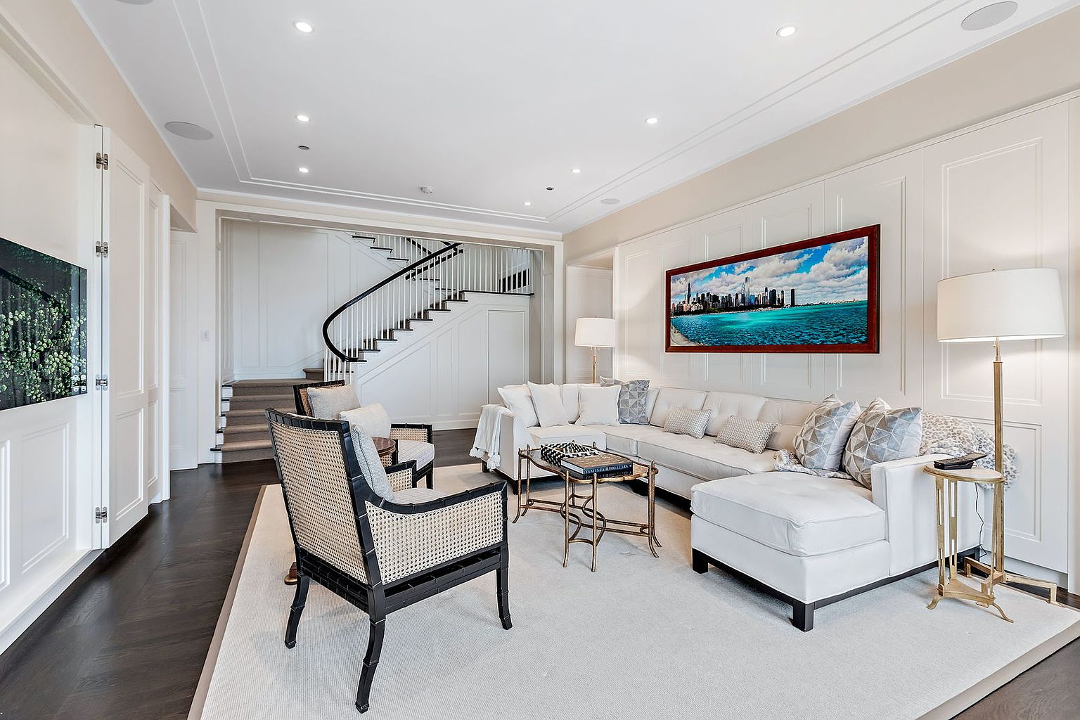 This is an elegant living room featuring a white sectional sofa, two cane-backed chairs, and a dark wood floor partially covered by a light-colored area rug. A large framed artwork hangs above the sofa, and a staircase is visible in the background. The room is well-lit with recessed lighting and lamps, creating a bright and inviting atmosphere.
