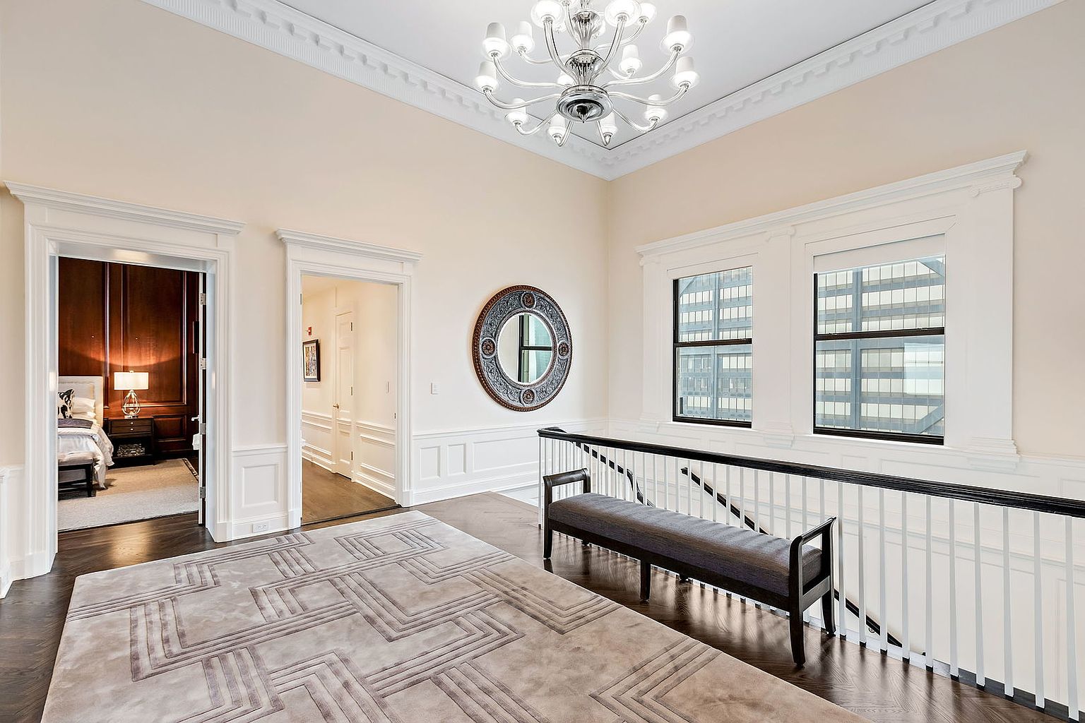 This interior shot showcases an elegant hallway with dark hardwood floors and a light-colored rug featuring a geometric pattern. The hallway features white wainscoting, a dark wood railing, and a bench, leading to doorways and windows that offer views of the city. A decorative mirror and a chandelier add to the sophisticated ambiance.
